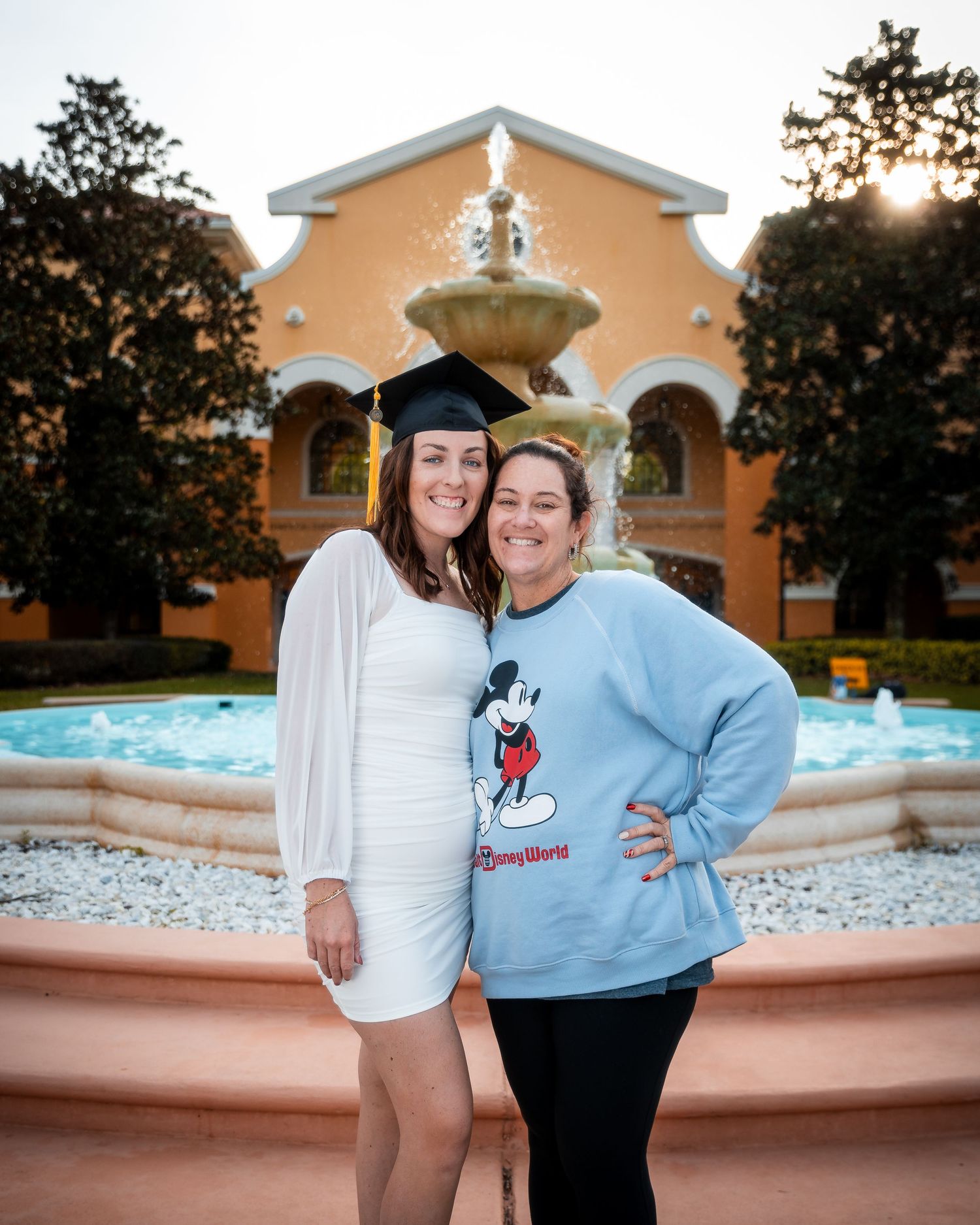Two people in casual attire share a joyful moment together outdoors near a beige building with a fountain.