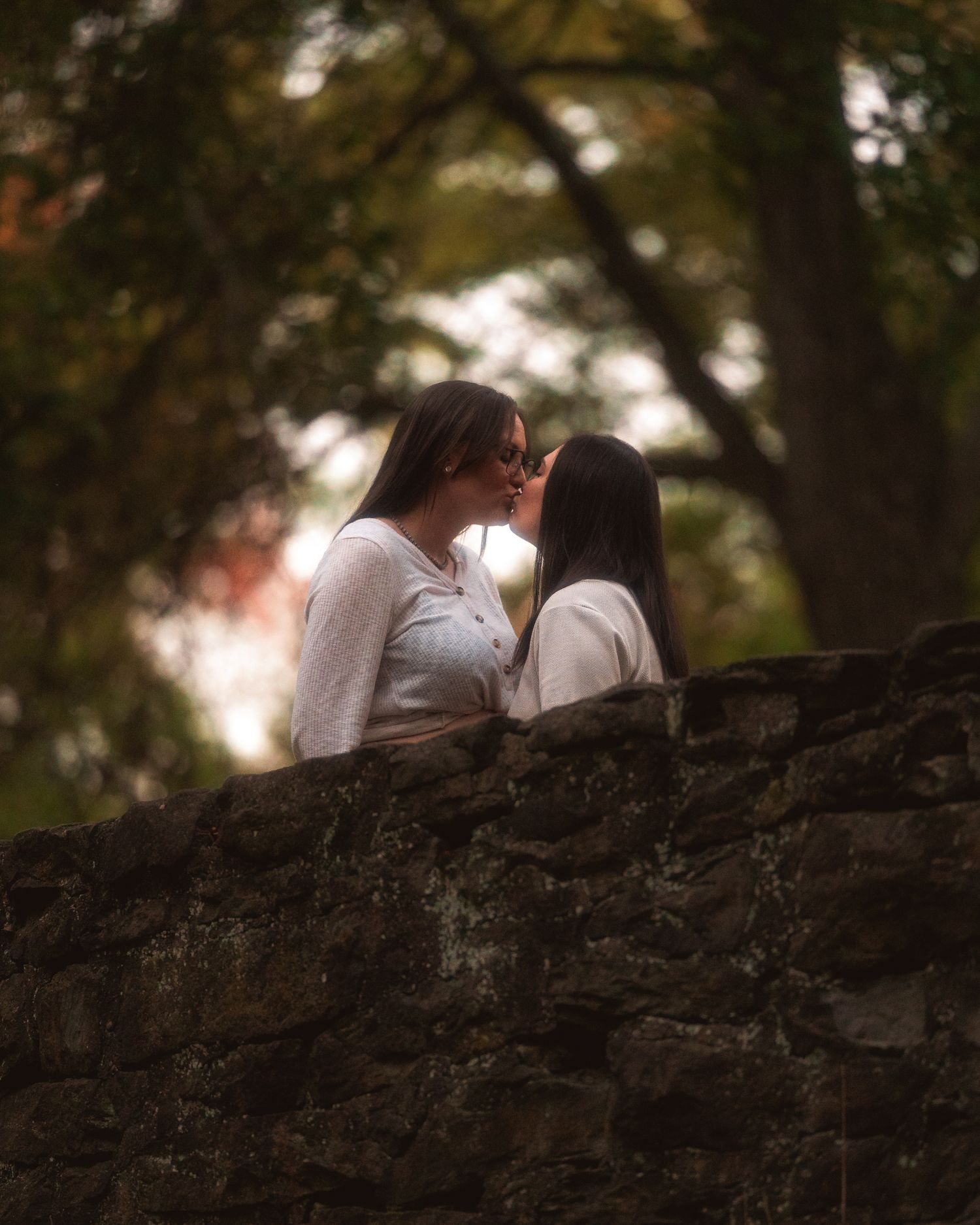 A couple sits intimately together on a stone wall in a wooded outdoor setting during golden hour.