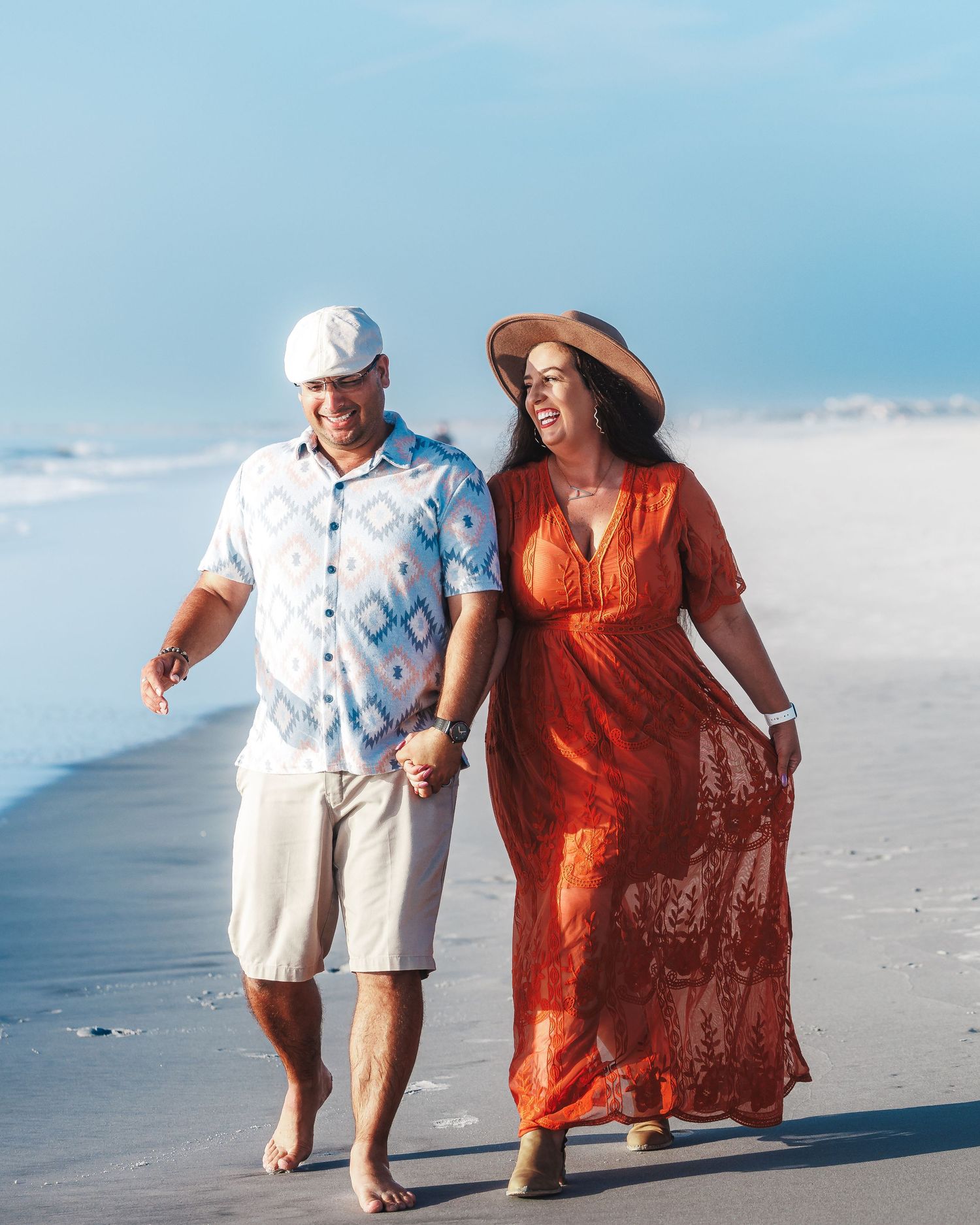 A couple walking along a beach shoreline with the person in orange dress flowing in the ocean breeze.