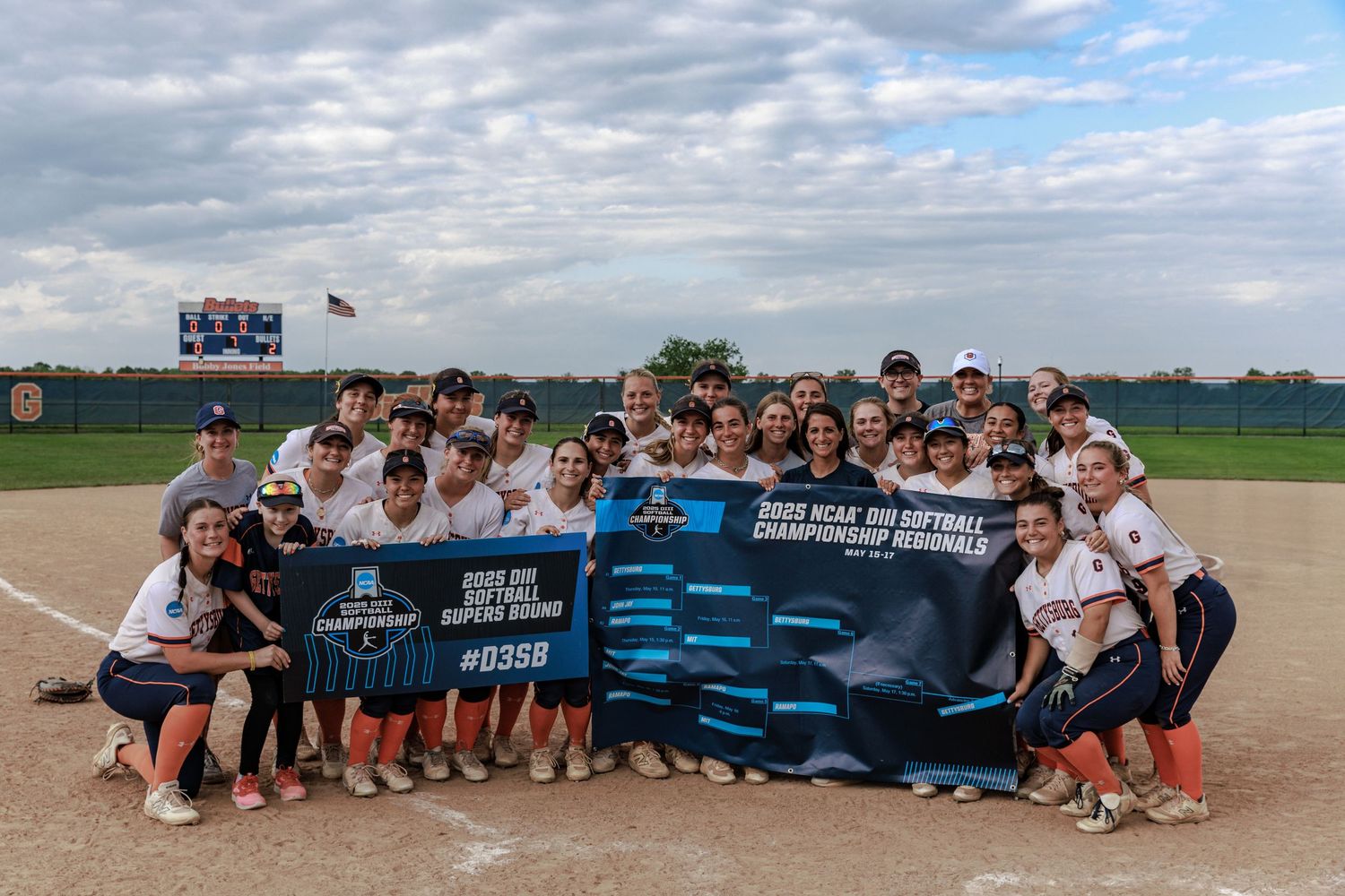 Large group of athletes pose together on a baseball field holding a blue banner under cloudy skies.