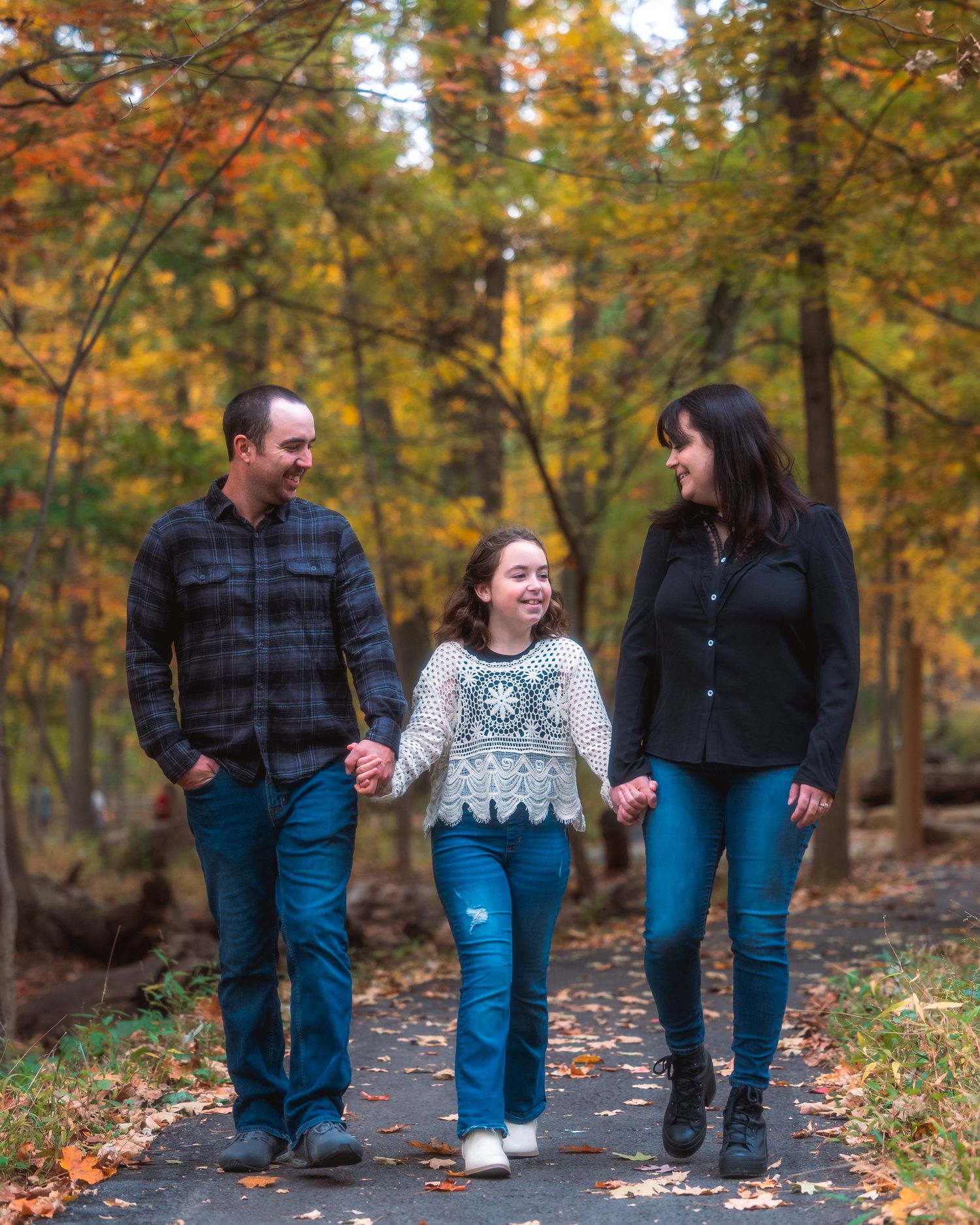 A couple walks hand in hand along a wooded path surrounded by vibrant autumn foliage and fallen leaves.