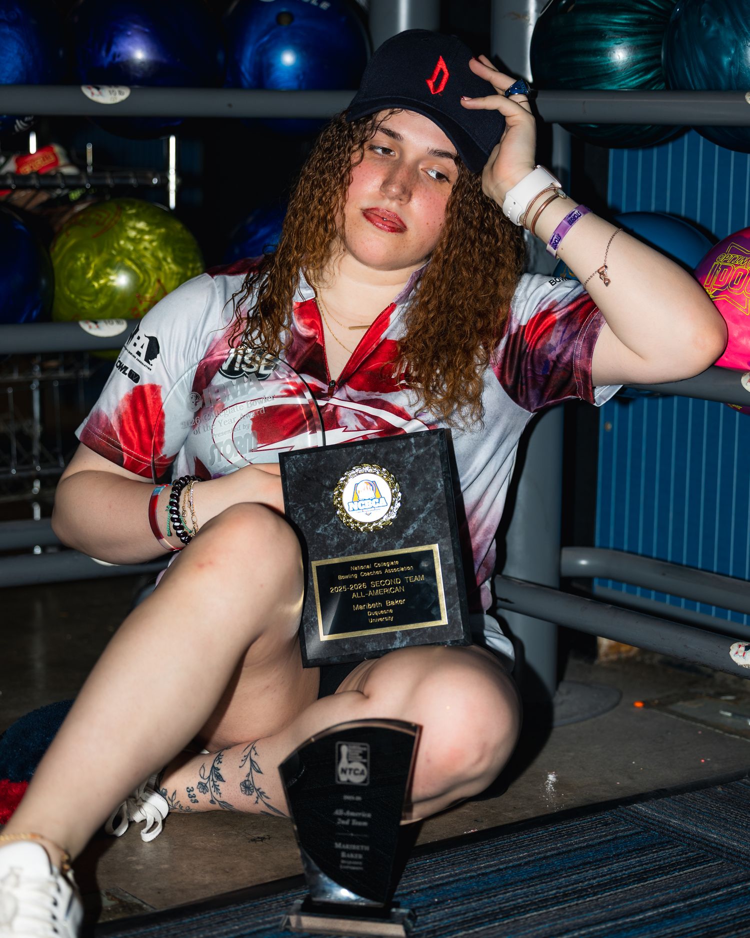 Young woman holding bowling trophy and plaque at a bowling alley, wearing a sports jersey and cap.