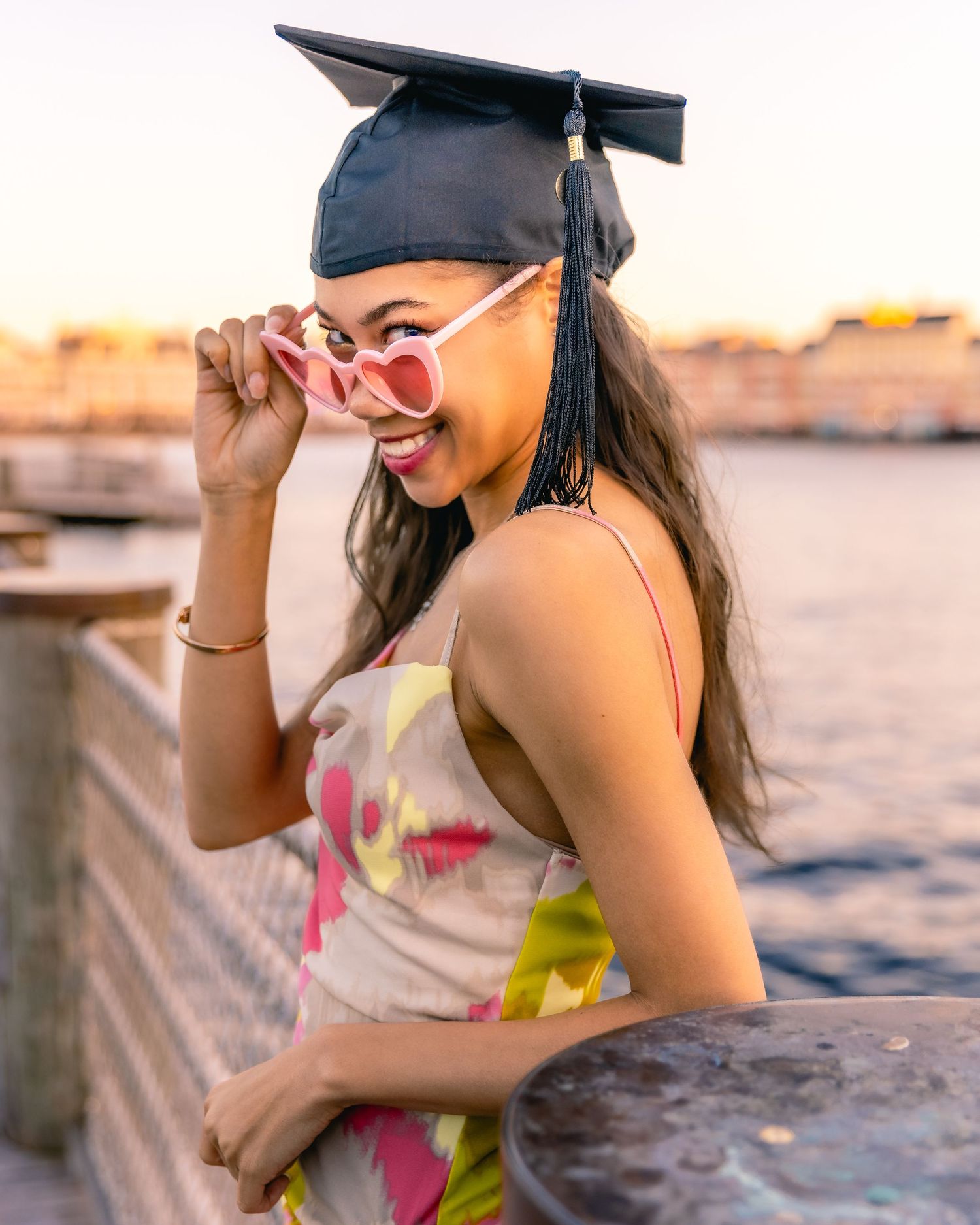Graduate in cap and gown wearing heart-shaped sunglasses poses by waterfront during golden sunset.