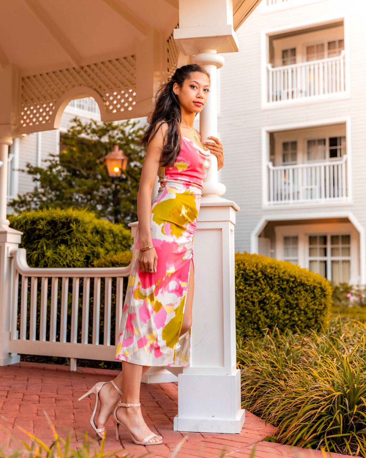 Woman in pink floral dress and heels poses gracefully on brick walkway at luxury resort property.
