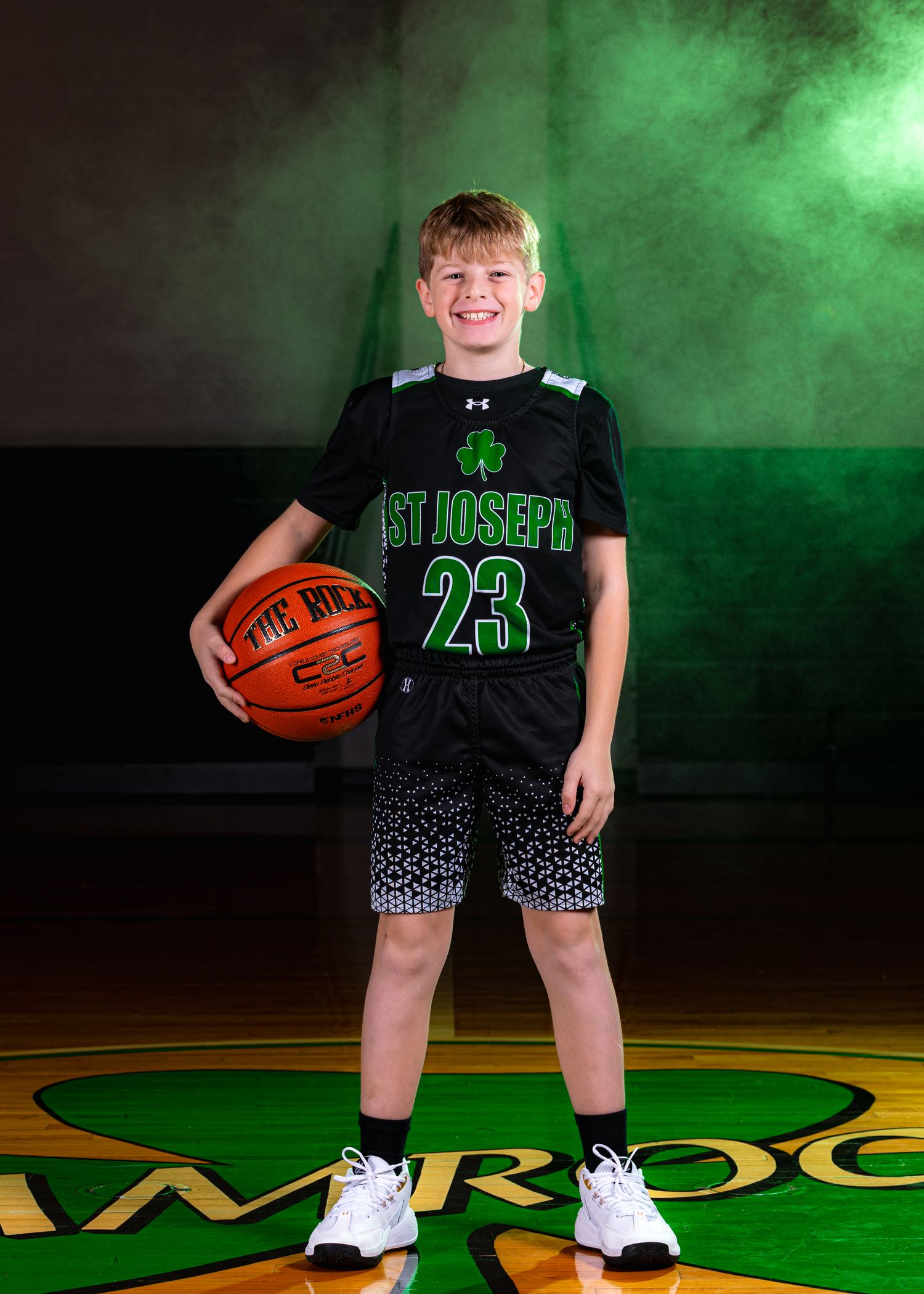 A basketball player in a dark green uniform poses with a ball against a smoky green backdrop for a sports photo.
