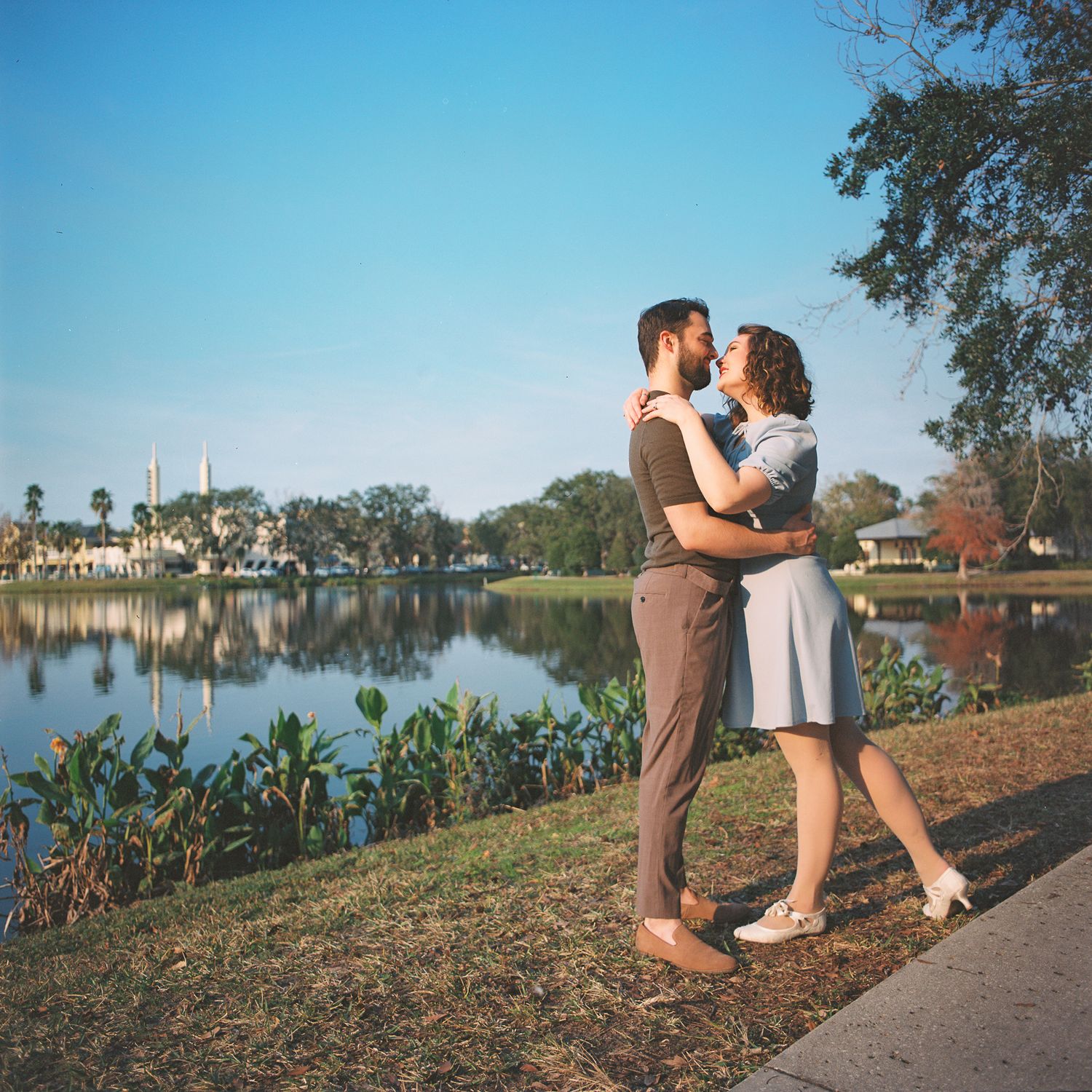 A couple shares a romantic moment by a serene lake with autumn foliage and reflective water at sunset.