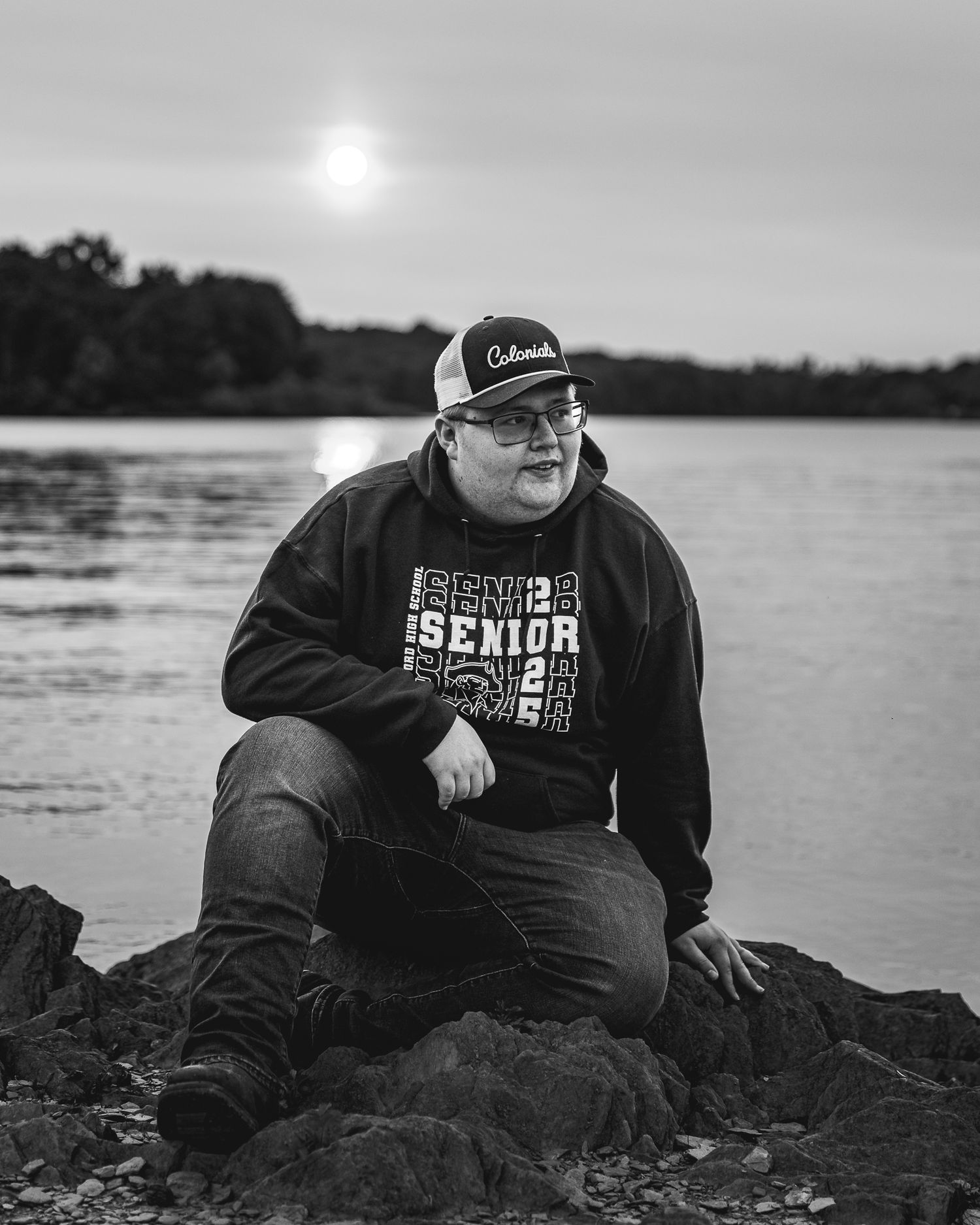 Black and white portrait of someone resting on rocky shoreline beside a lake at dusk.