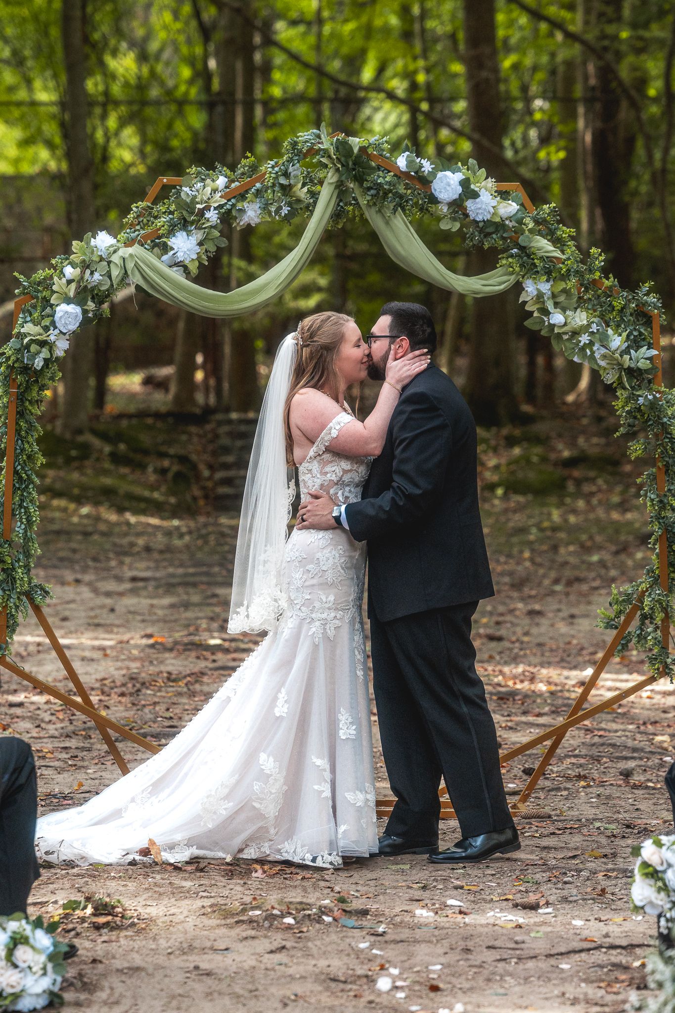 Wedding couple shares a romantic kiss under a floral arch decorated with blue and white flowers in a forest setting.