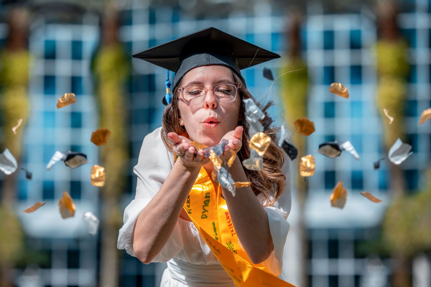 Graduate in cap and gown blowing glitter confetti in sunlight during outdoor celebration ceremony.
