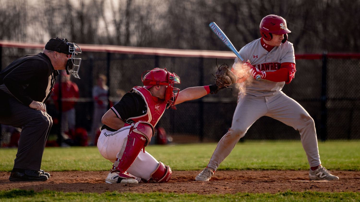 Baseball batter in white uniform at plate with catcher in red and umpire during evening game.