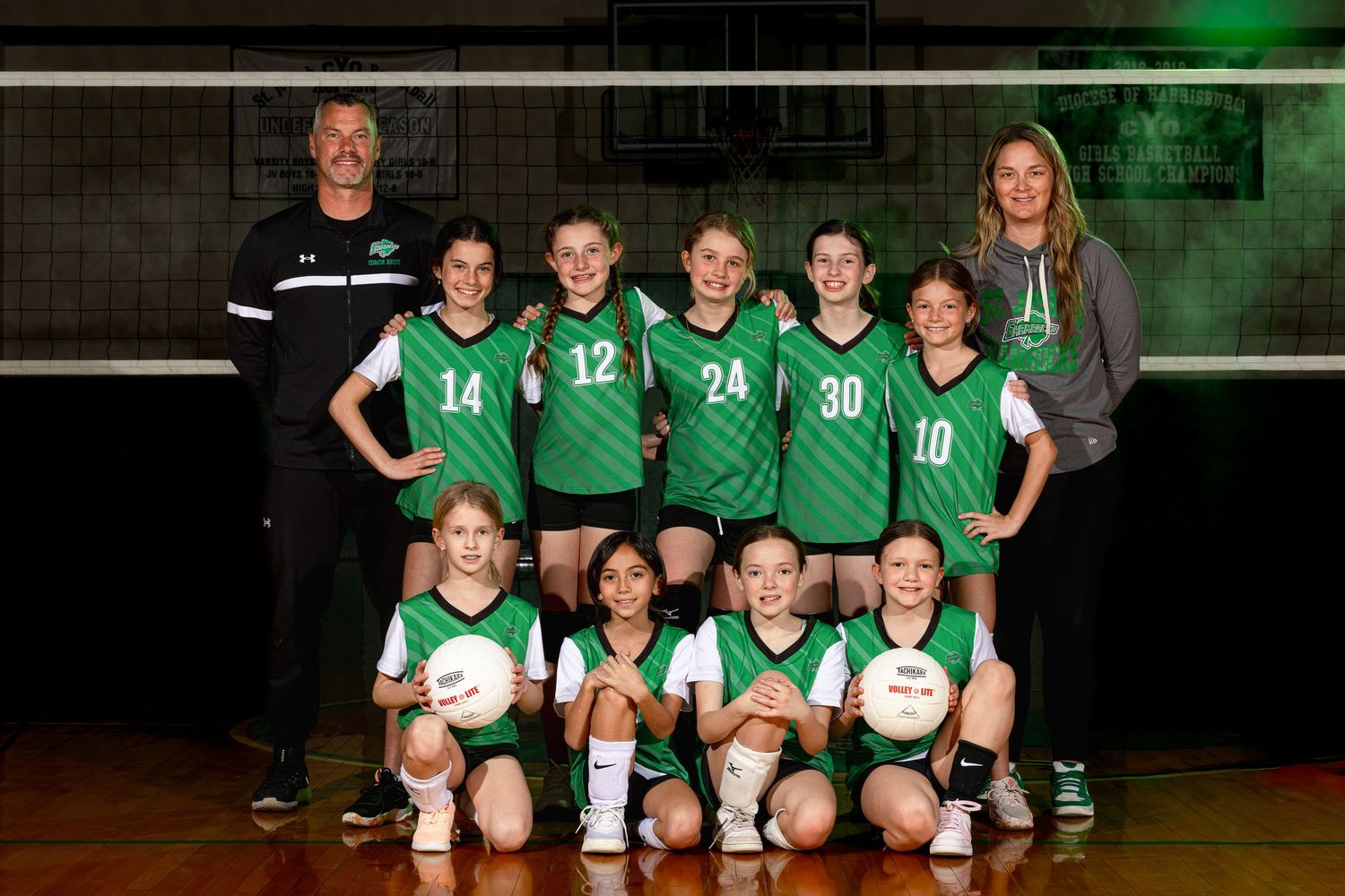 Youth girls volleyball team in green jerseys posing for team photo with two coaches in a gymnasium.