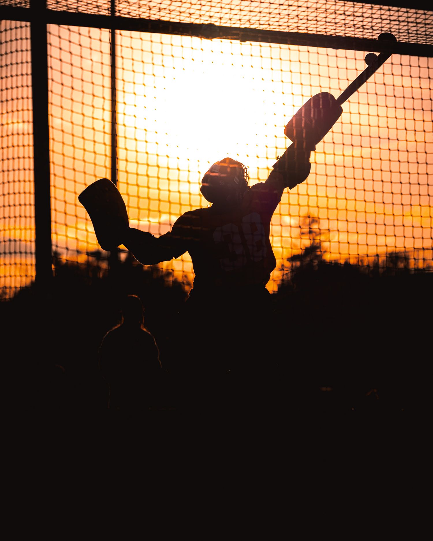 Silhouettes of people playing baseball against a dramatic orange sunset sky with protective netting in view.