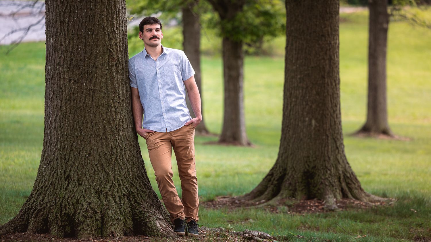 A person in casual attire leans against a large tree trunk in a peaceful park setting on a sunny day.