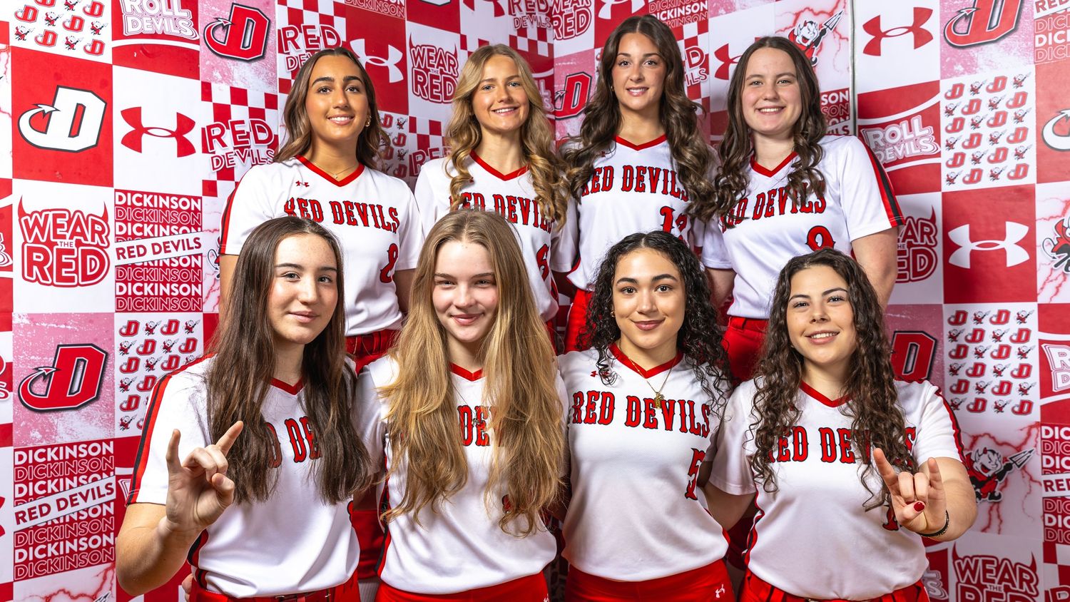 Seven Red Devils volleyball players pose in white uniforms in front of a red and white branded backdrop.