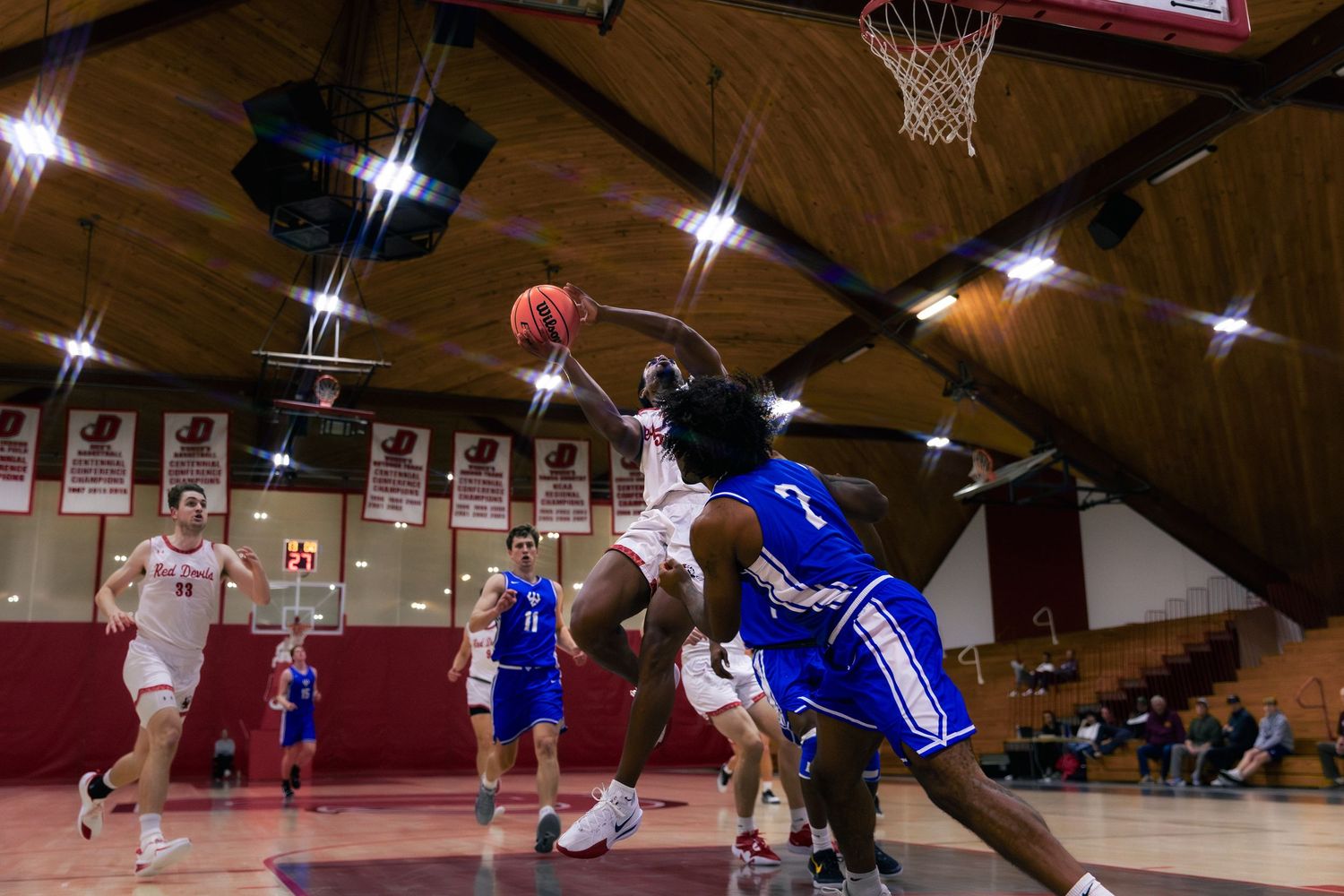 Basketball players compete for a rebound during an intense indoor game with dramatic lighting in a gymnasium.