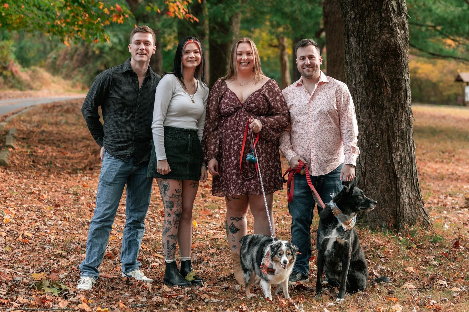 A group of four people and two dogs pose together on a path covered in fallen autumn leaves in a park setting.