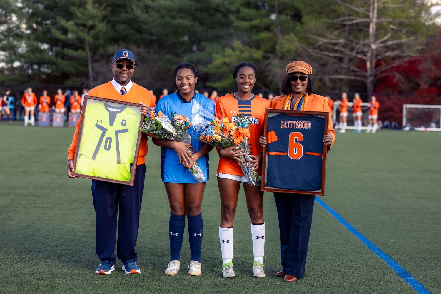 Youth sports team players and coaches line up on a grass field wearing orange and blue uniforms.