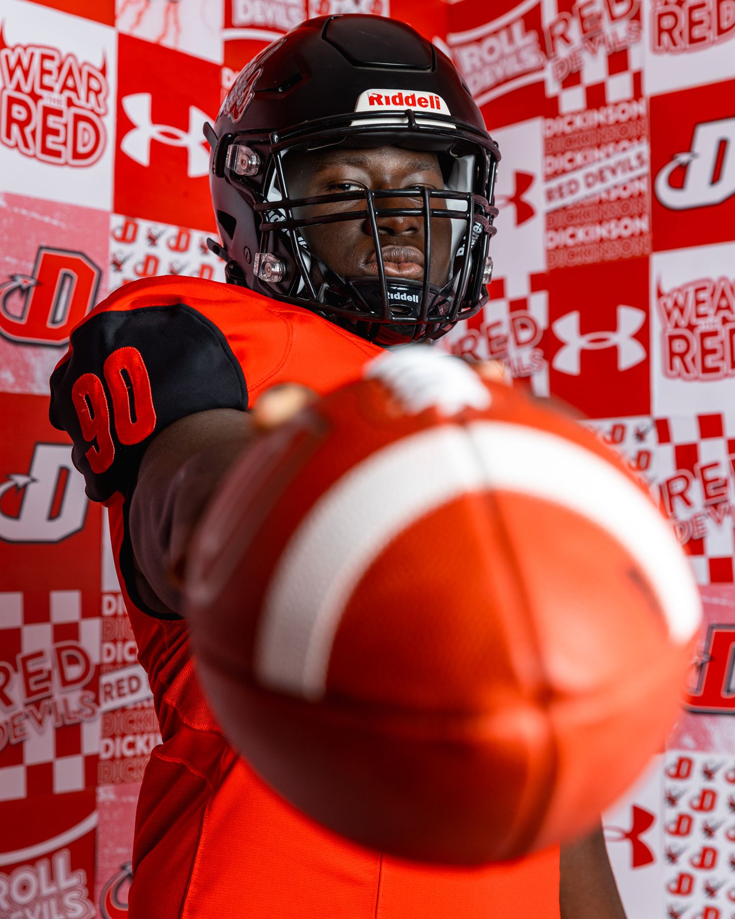 Football player in Dickinson Red Devils #90 orange jersey holding a football toward camera.