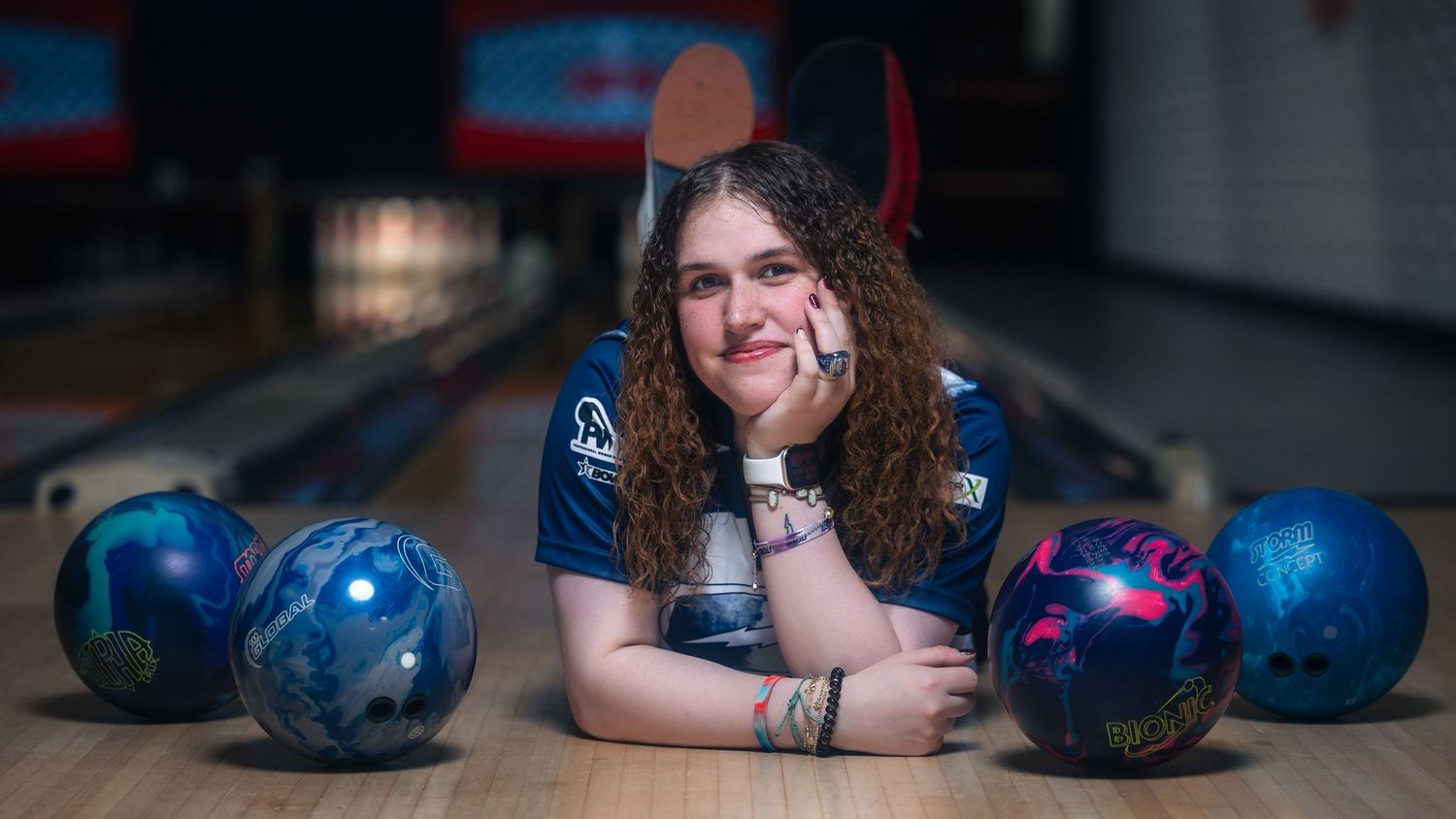 Young female bowler poses on lane surrounded by colorful bowling balls in a bowling alley.