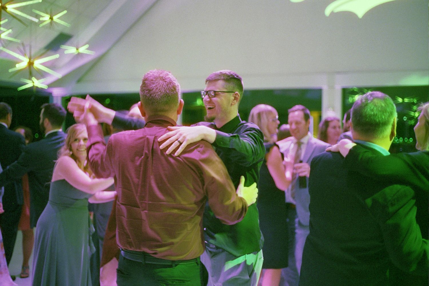 Couples dance together in a dimly lit room with green and pink lighting creating an atmospheric ambiance.