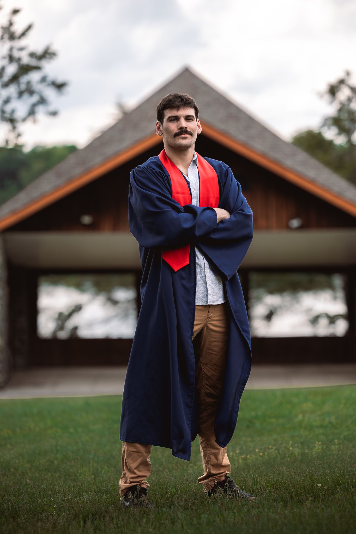 A person in graduation attire stands on grass in front of wooden buildings on an overcast day.