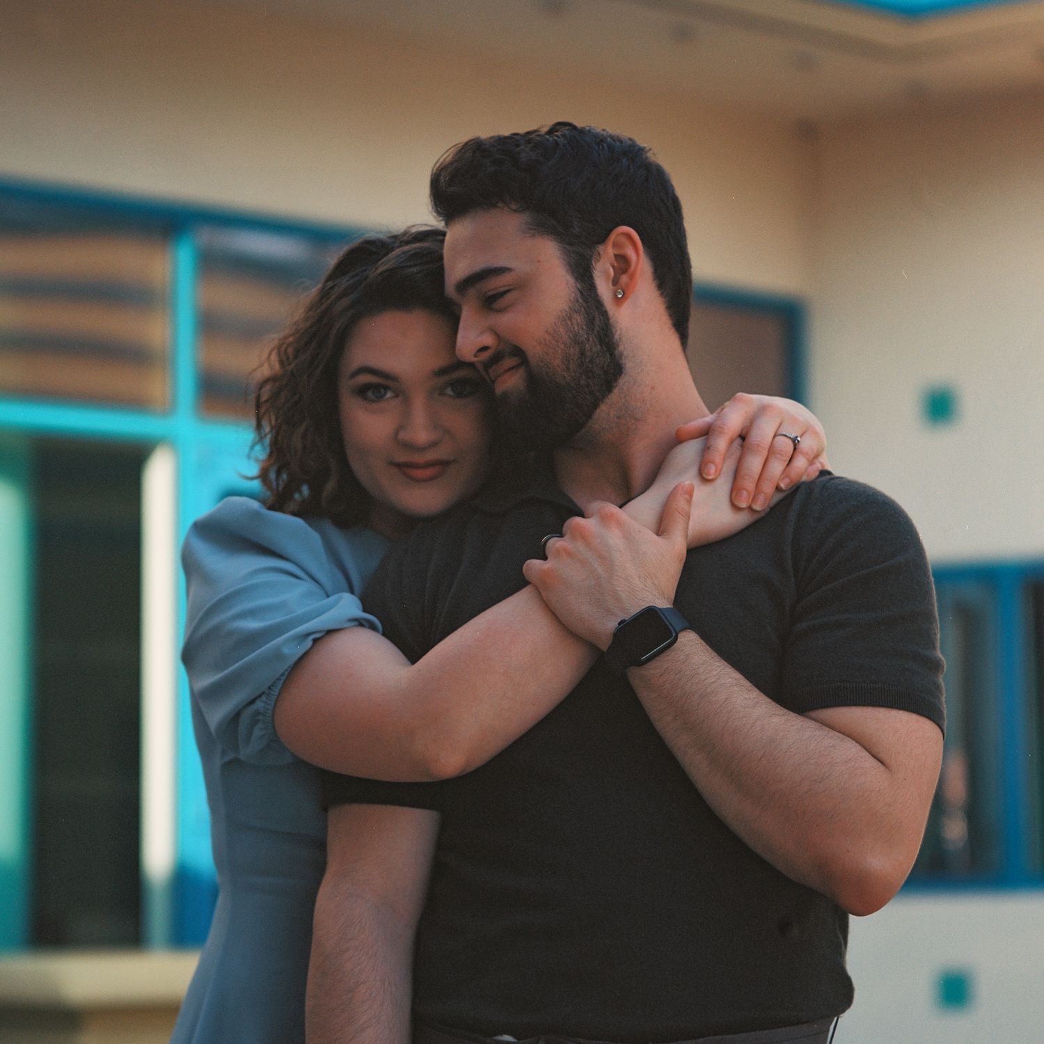 A couple in dark clothing embraces in a dance-like pose against a light colored interior background.