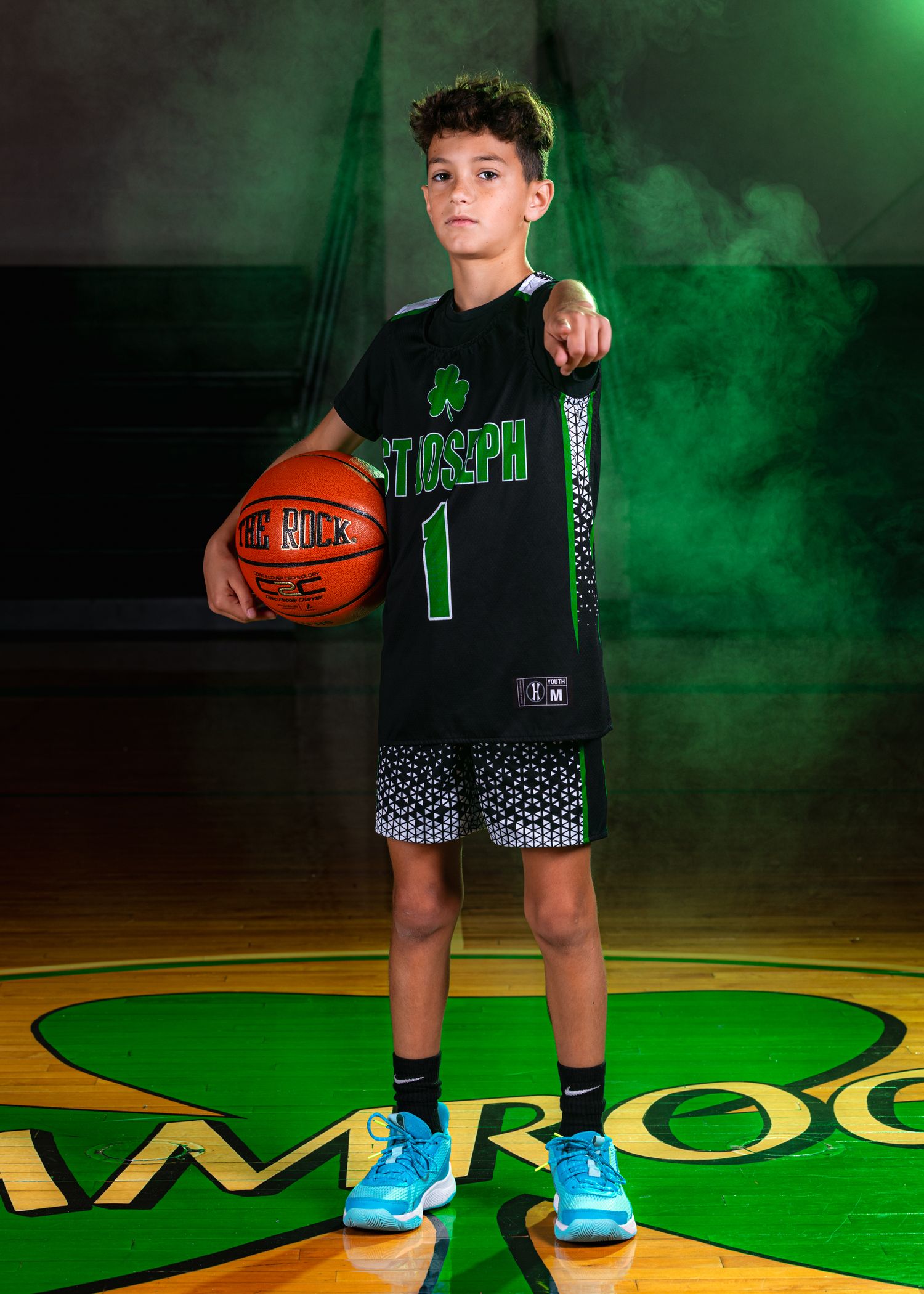 Basketball player in black uniform and blue shoes poses against green smoke effect on wooden court.