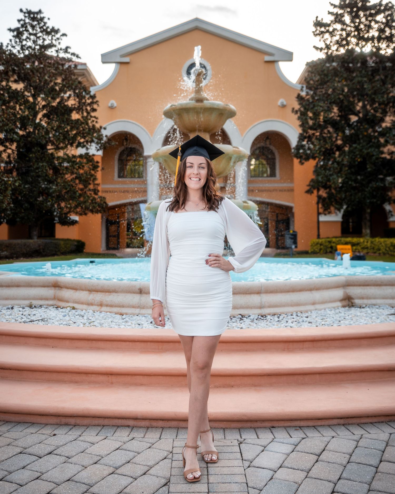 Graduate in white dress and cap poses by fountain at sunset on campus.