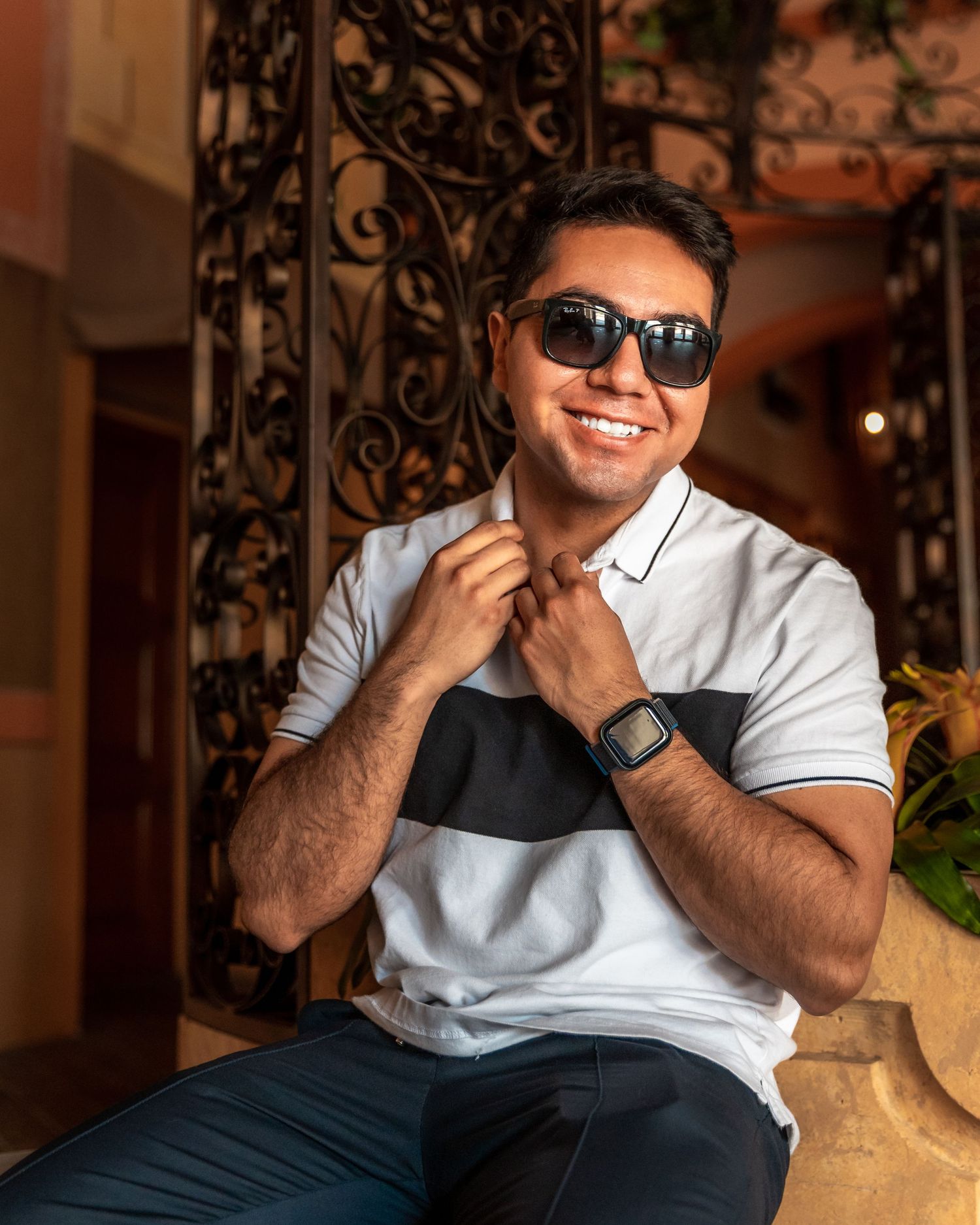 Person in sunglasses and white polo shirt with black stripe smiles while sitting by decorative iron gate.