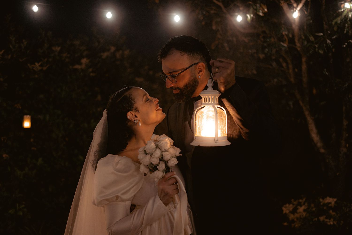 A romantic nighttime wedding scene with a couple illuminated by a vintage lantern among trees.