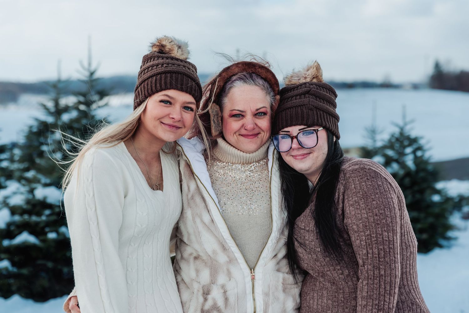 Three friends wearing cozy winter sweaters and knit hats smile together in a snowy landscape with evergreen trees.