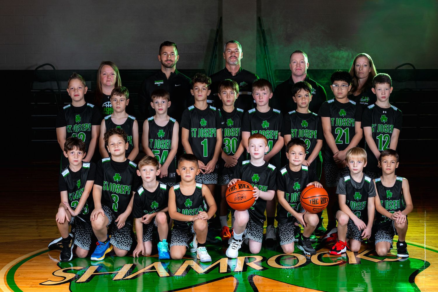 Youth basketball team members pose in black uniforms on a wooden court with green lighting in the background.