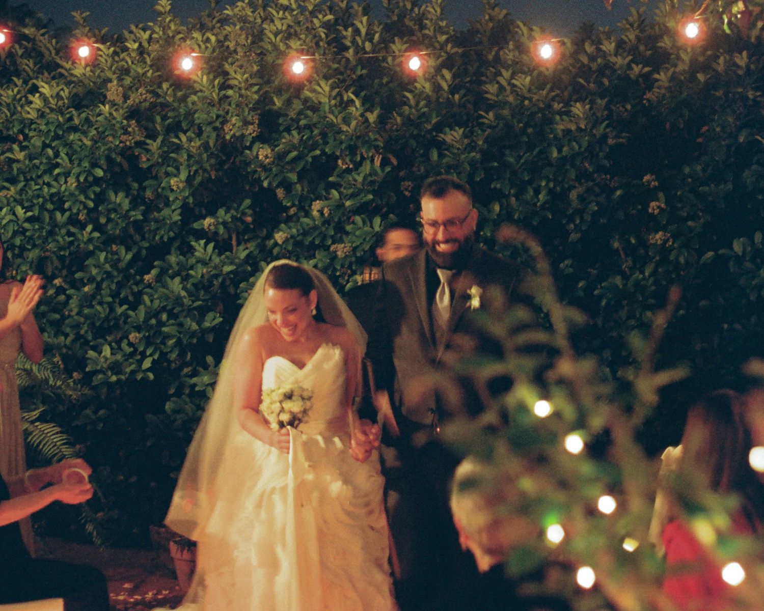 A couple dances together at an outdoor evening wedding reception with string lights glowing in the background.