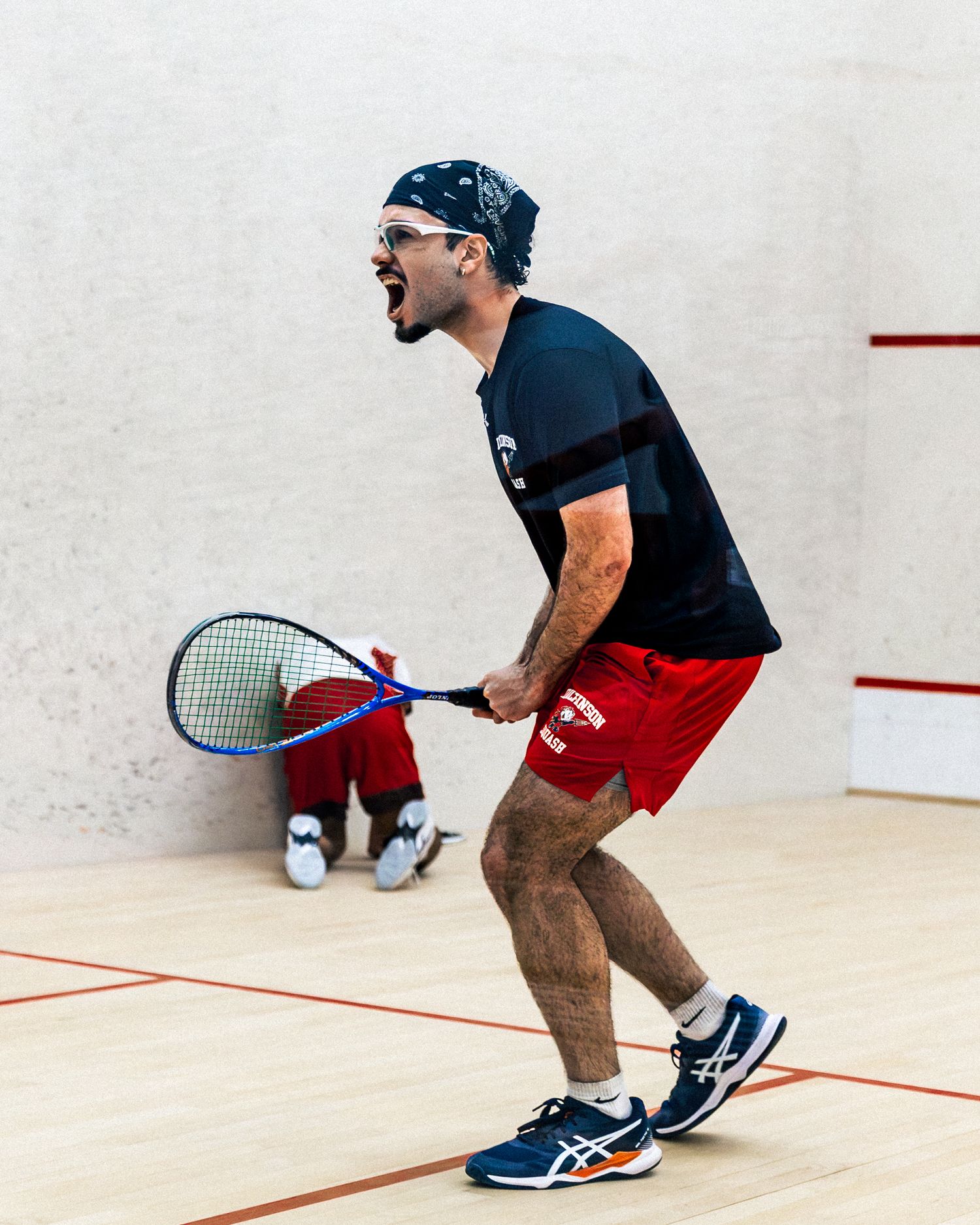 A squash player in black shirt and red shorts shows intense focus during a match on an indoor court.