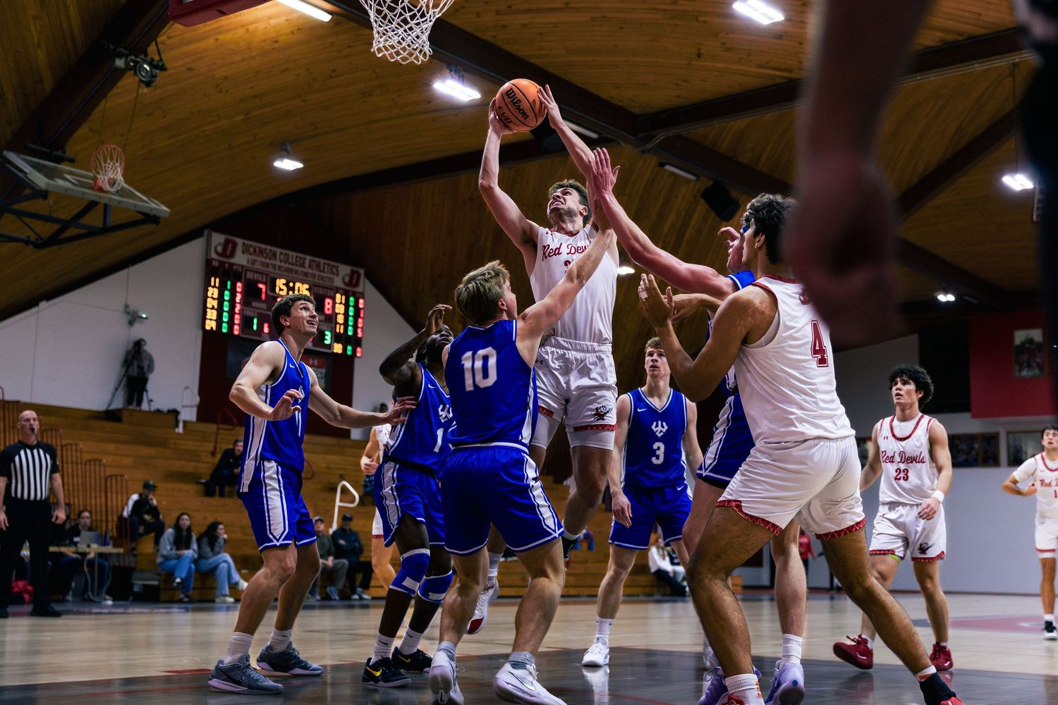 High school basketball players compete for a rebound during an indoor game, with players in white and blue uniforms.