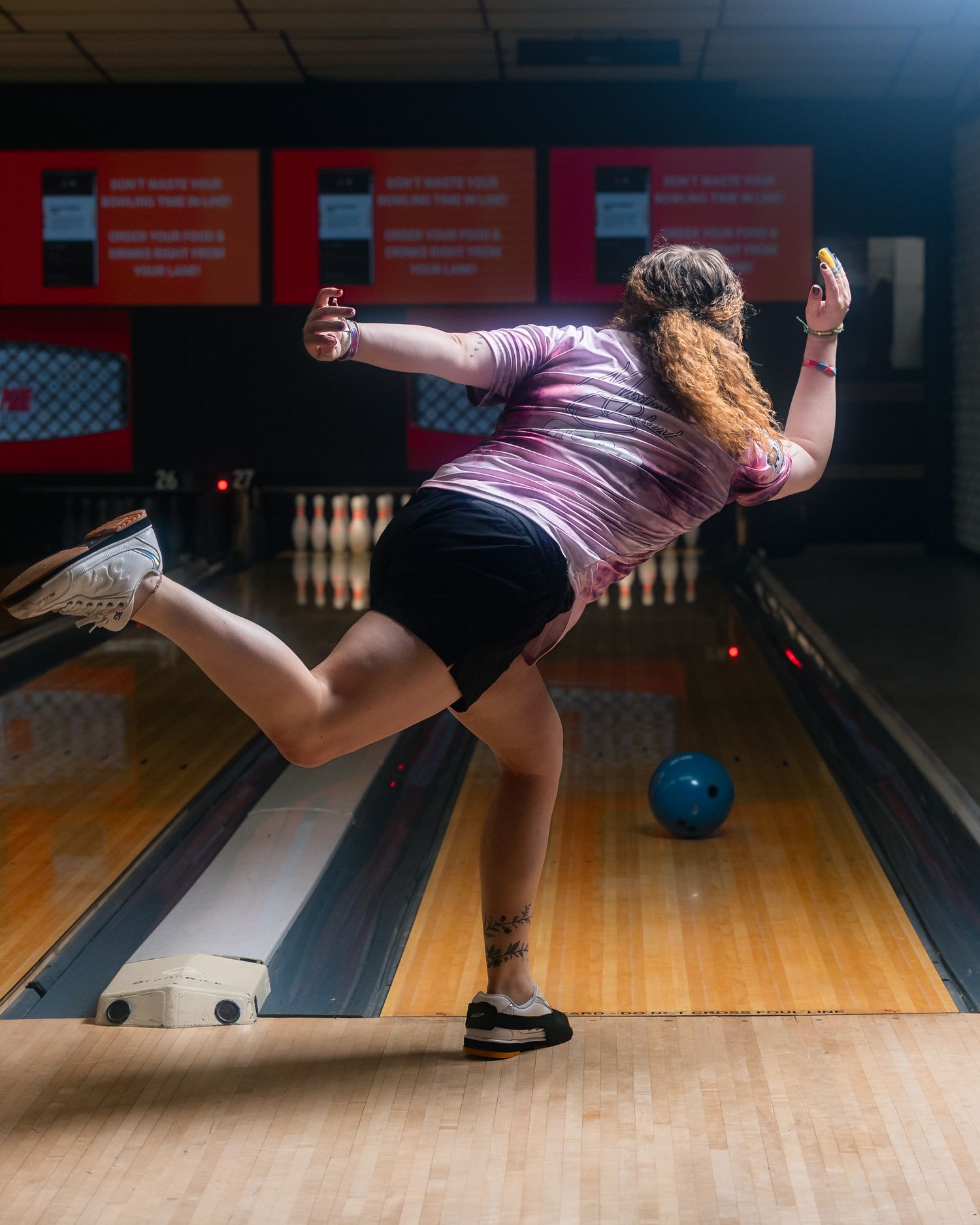 Woman in pink shirt releases bowling ball on lane, mid-throw with leg raised, pins visible in background.