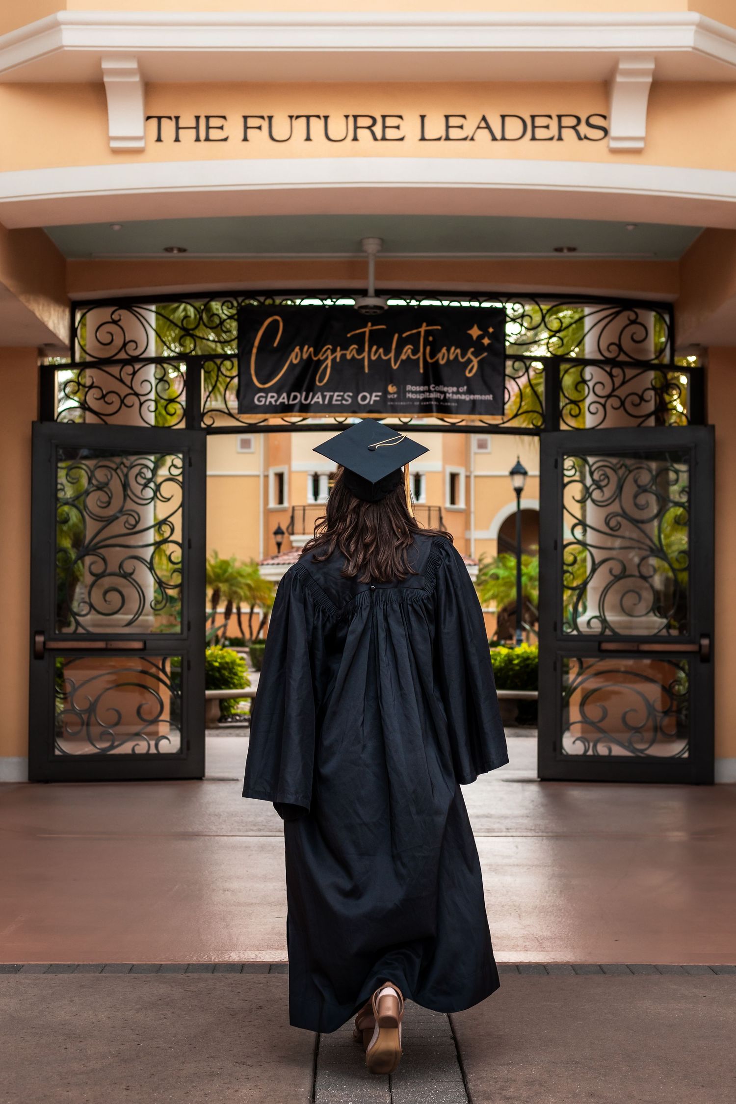 Graduate poses in black cap and gown in front of building with 'The Future Leaders' sign.