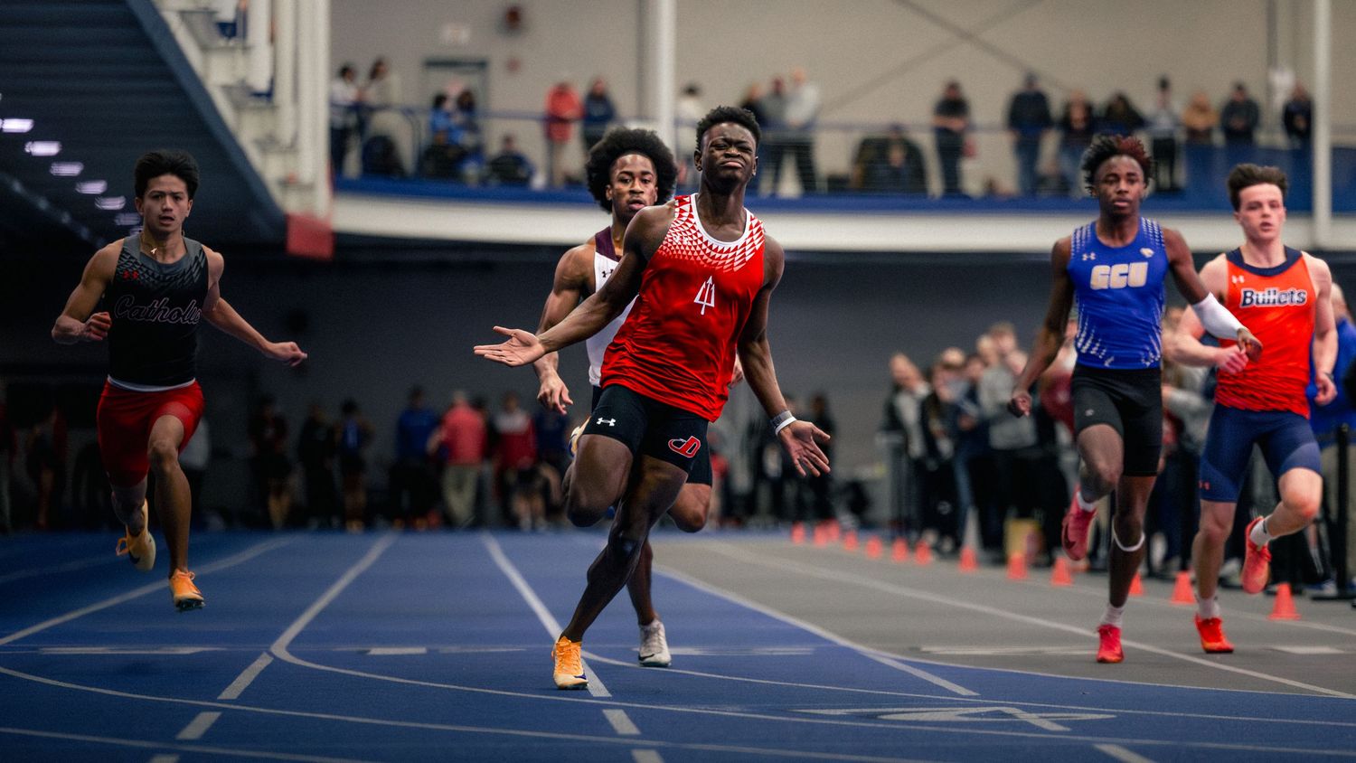 Athletes compete in a high-speed indoor track race with multiple runners in close formation.