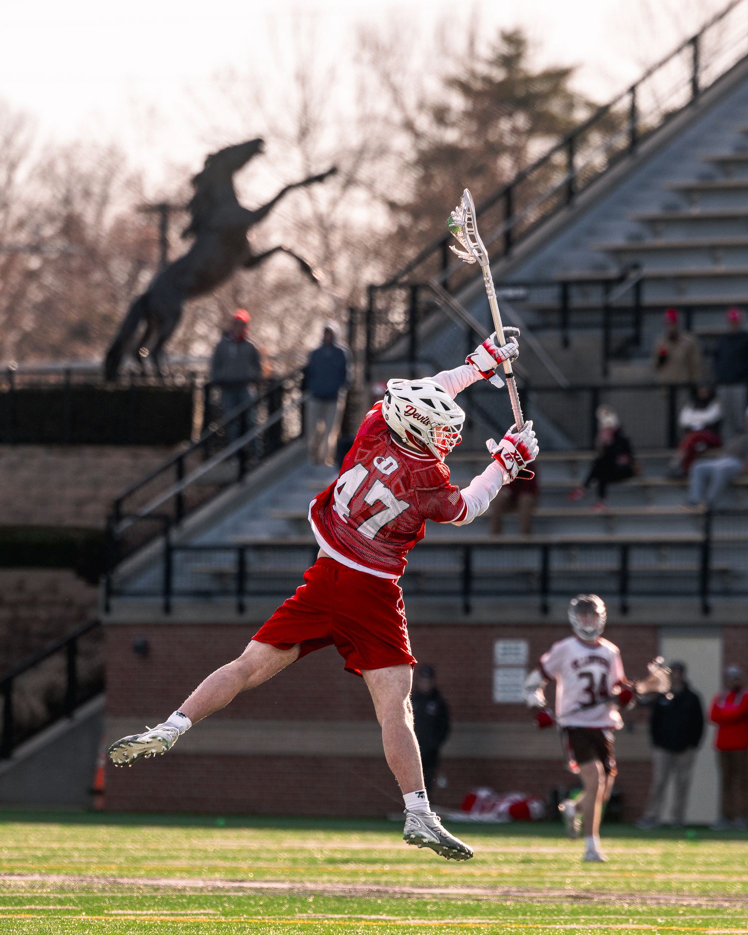 A lacrosse goalie makes a diving save during a match, stretching horizontally in mid-air to block the shot.