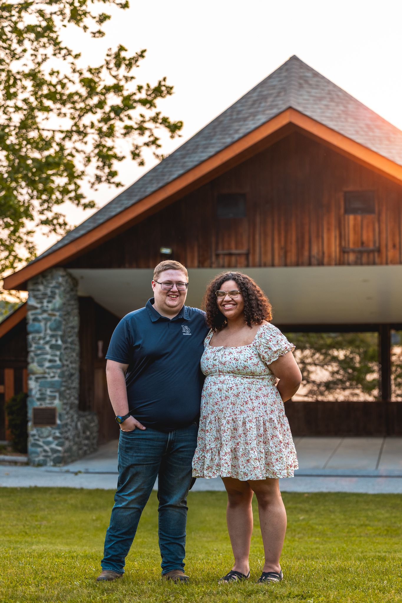 A couple poses together outdoors in front of a rustic building with stone accents at sunset.