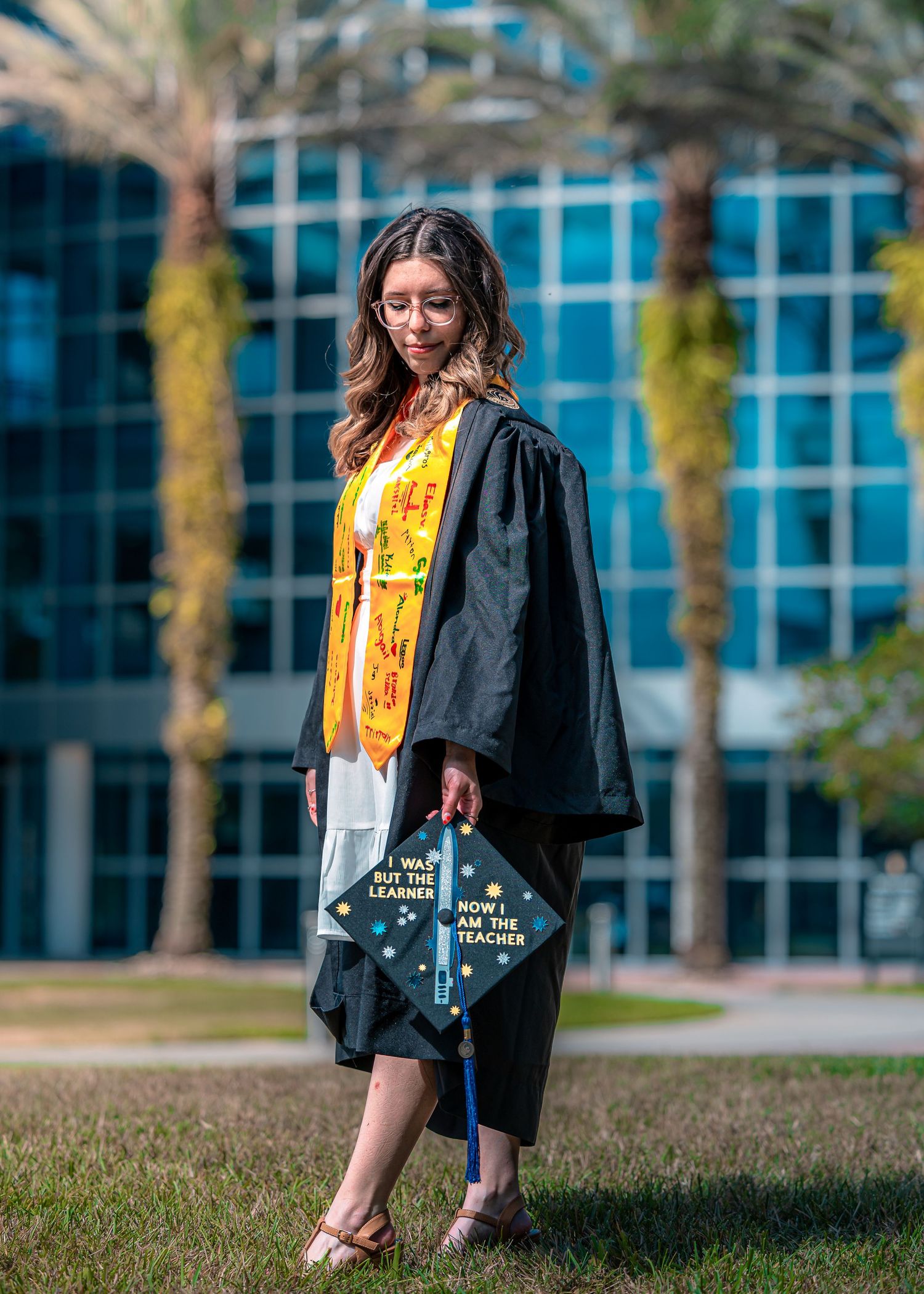 Graduate in black gown holds graduation cap decorated with stars and sparkles against modern campus building.