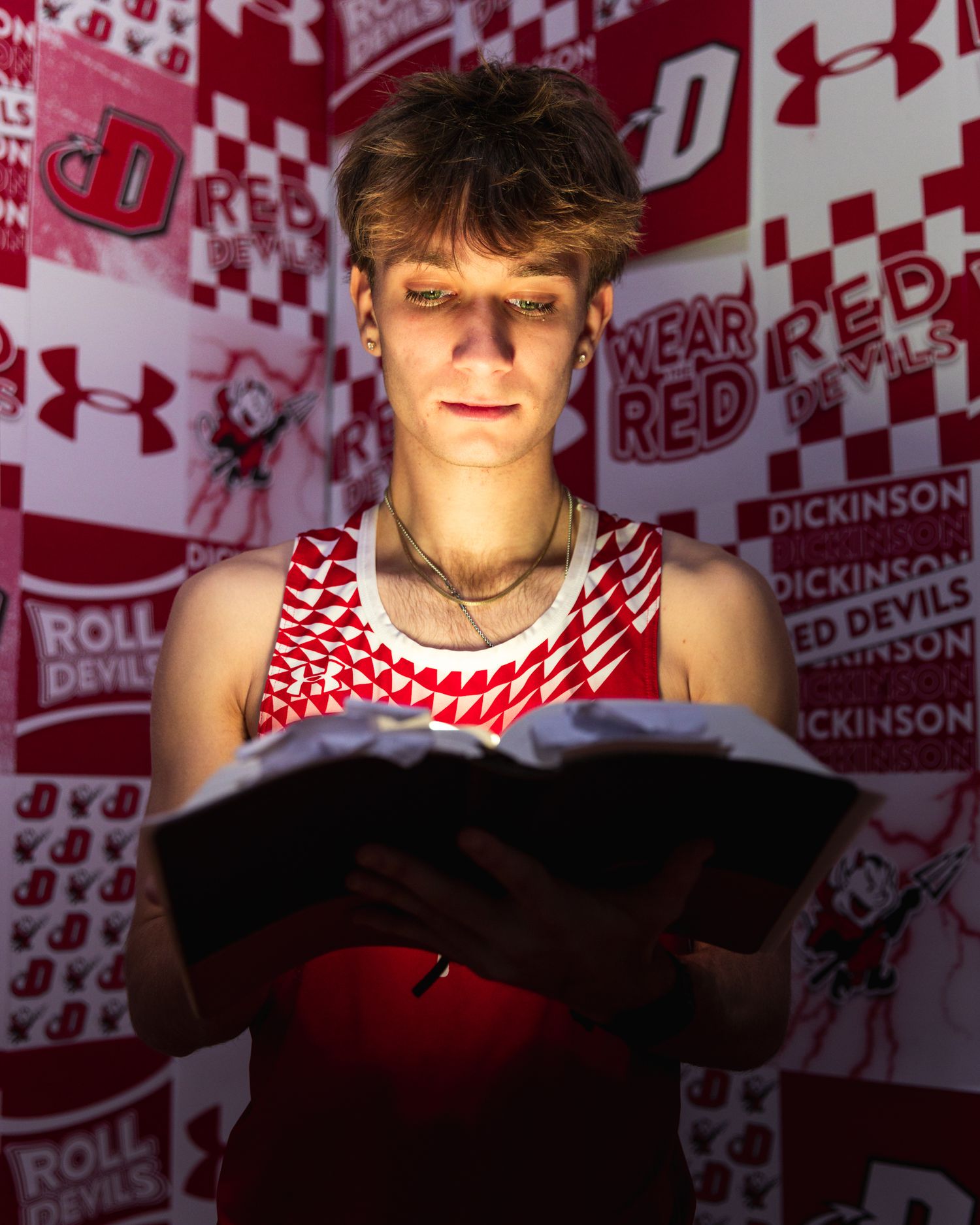 Teen athlete in red jersey reads book in front of Dickinson Red Devils themed backdrop with red and white logos.