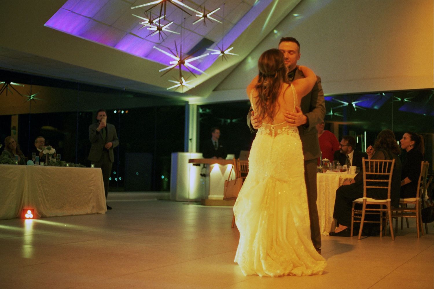A couple dances in a dimly lit reception hall with purple uplighting and white ceiling decorations.
