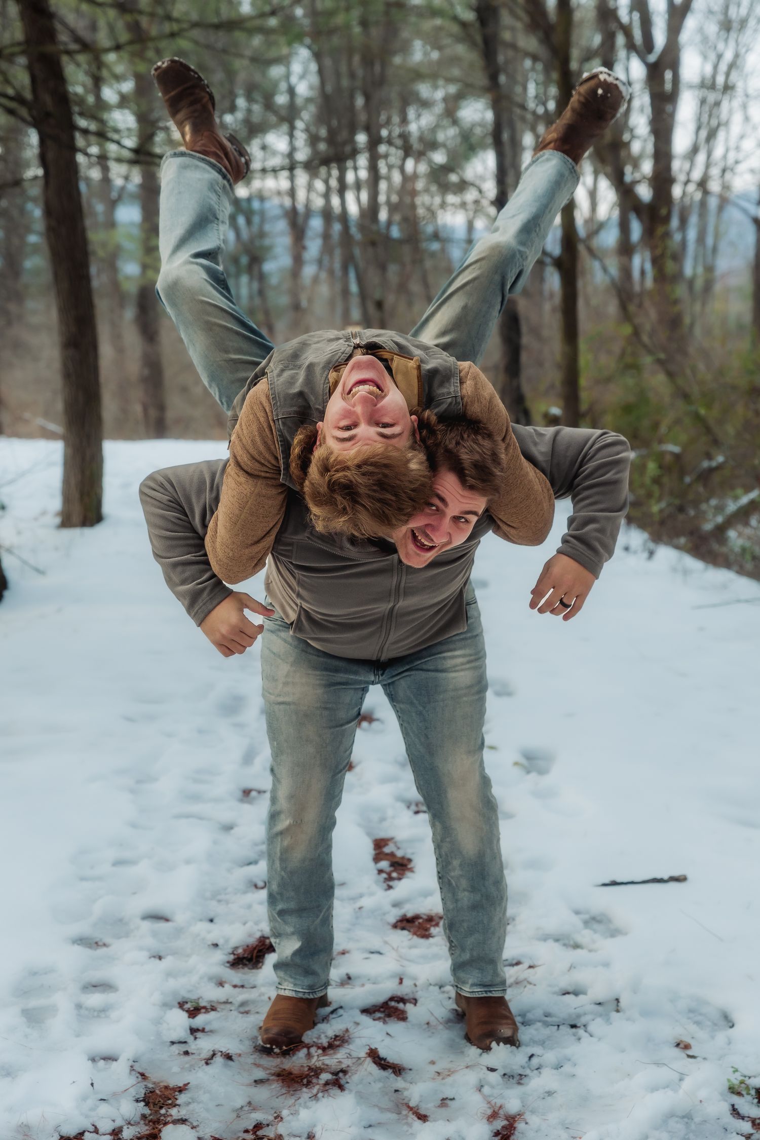 Two people playfully pose in a snowy winter forest scene with one person carrying the other upside down.