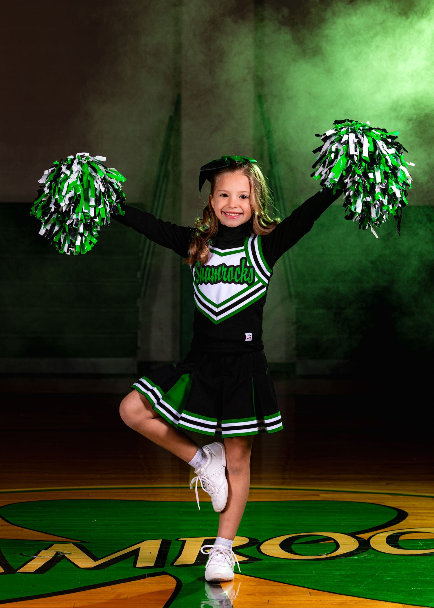 A cheerleader in green and white uniform performs dynamic poses and jumps on a basketball court with green lighting.