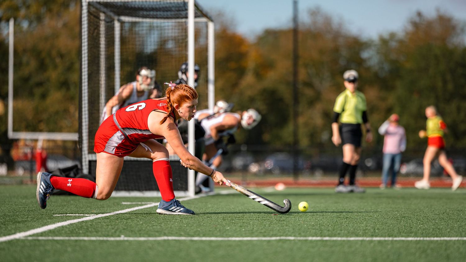 Field hockey player in red uniform takes a shot on goal during outdoor match on grass field.