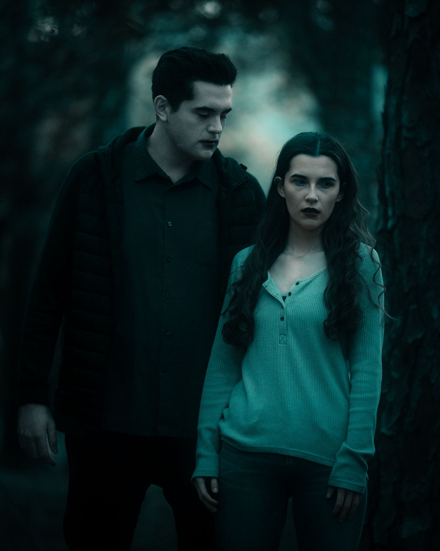 A dark and moody scene shows two figures in a misty forest with a blue-green cinematic color palette.