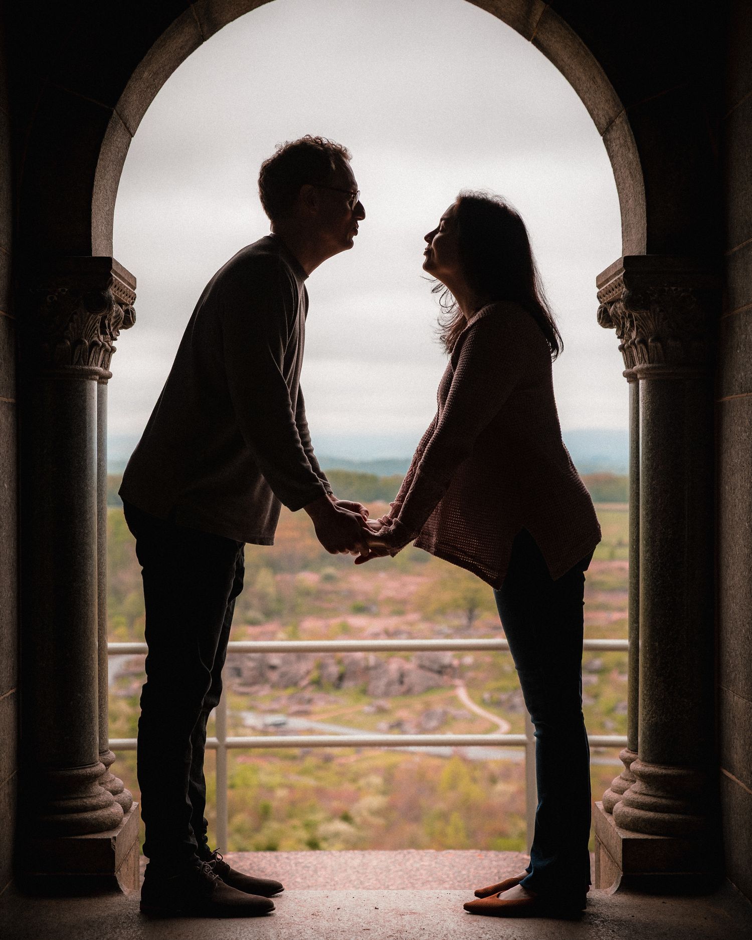 Romantic couple silhouetted in stone archway holding hands with scenic landscape backdrop.
