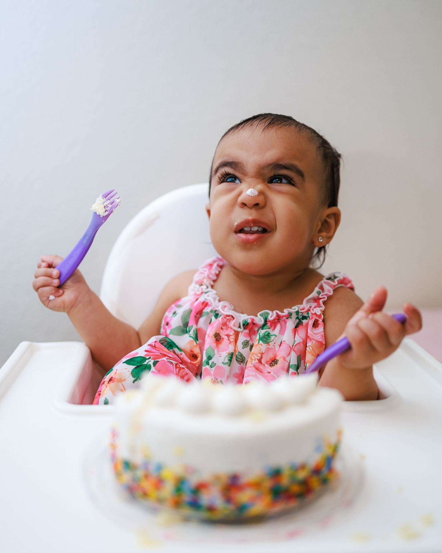 Child in floral dress sits in high chair with birthday cake decorated with colorful sprinkles and holds purple utensil.