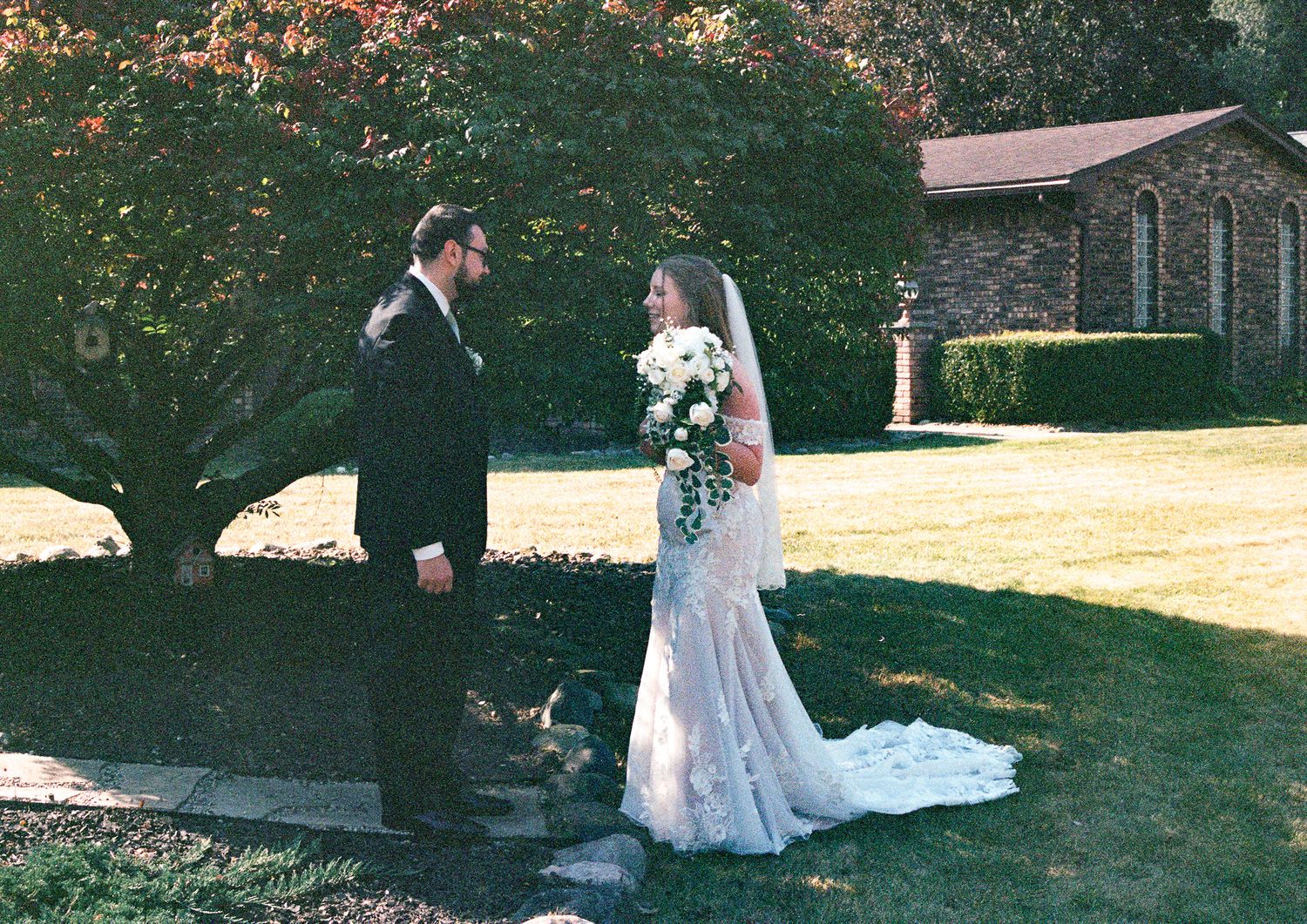 A wedding ceremony takes place under a large tree in a garden setting with guests seated on white chairs.